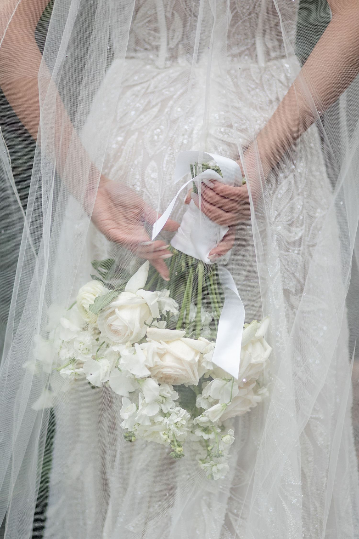 Bride in beaded gown holds white rose bouquet with ribbon behind her back.
