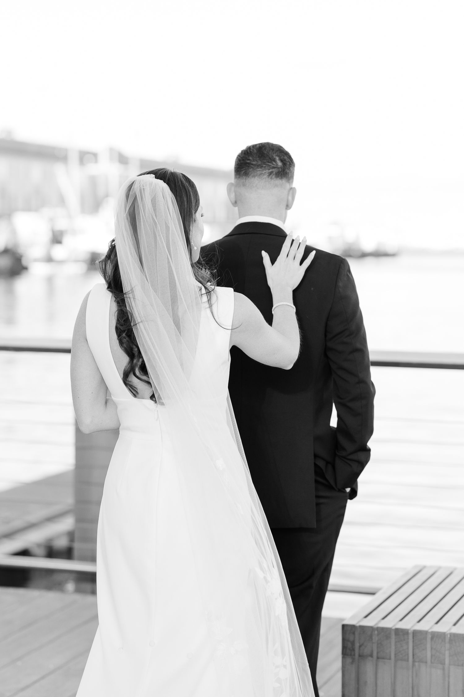 Black and white photo of bride and groom walking together toward marina, bride resting hand on groom.