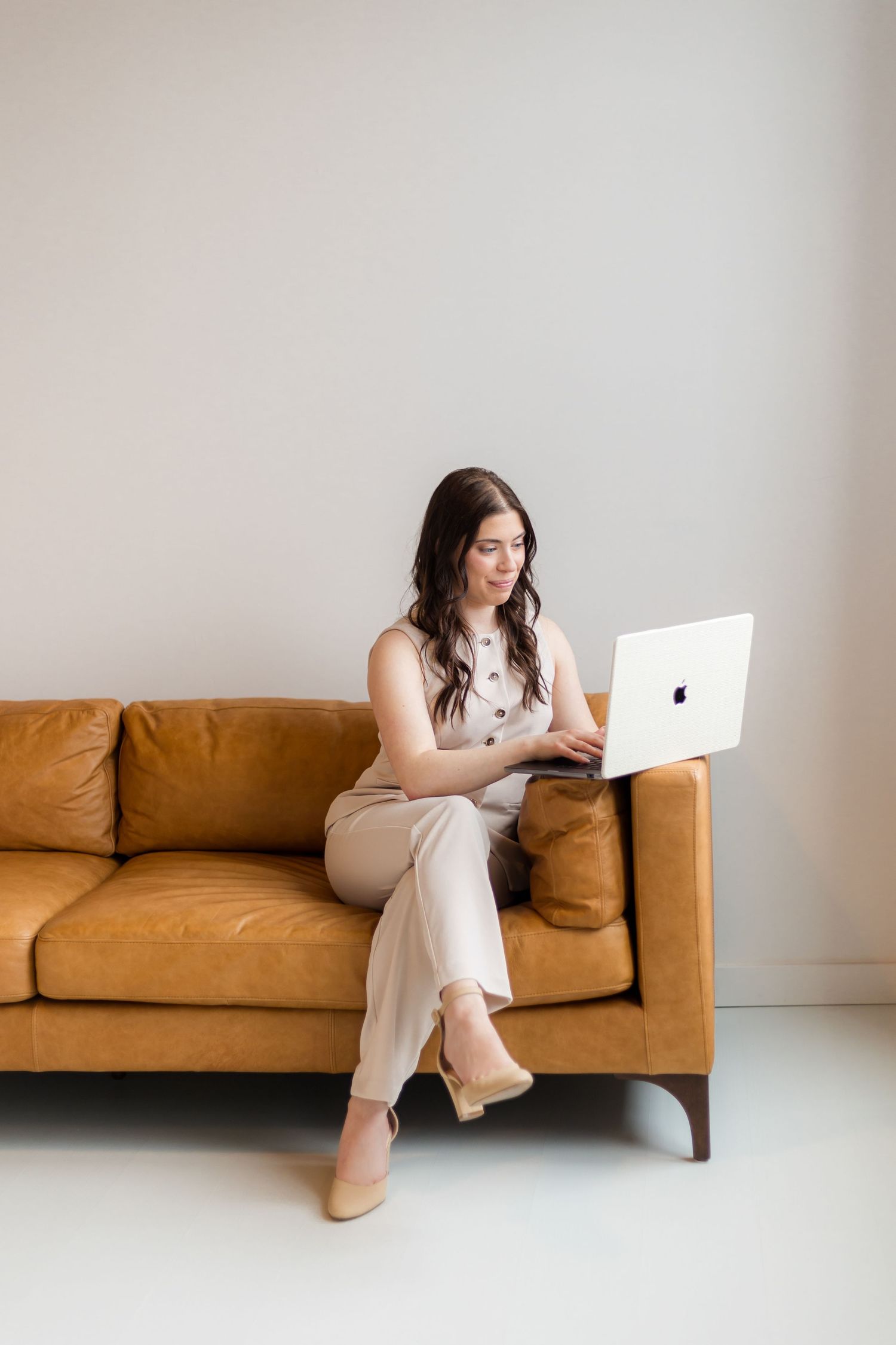 Professional woman in beige jumpsuit working on laptop while sitting on tan leather sofa.
