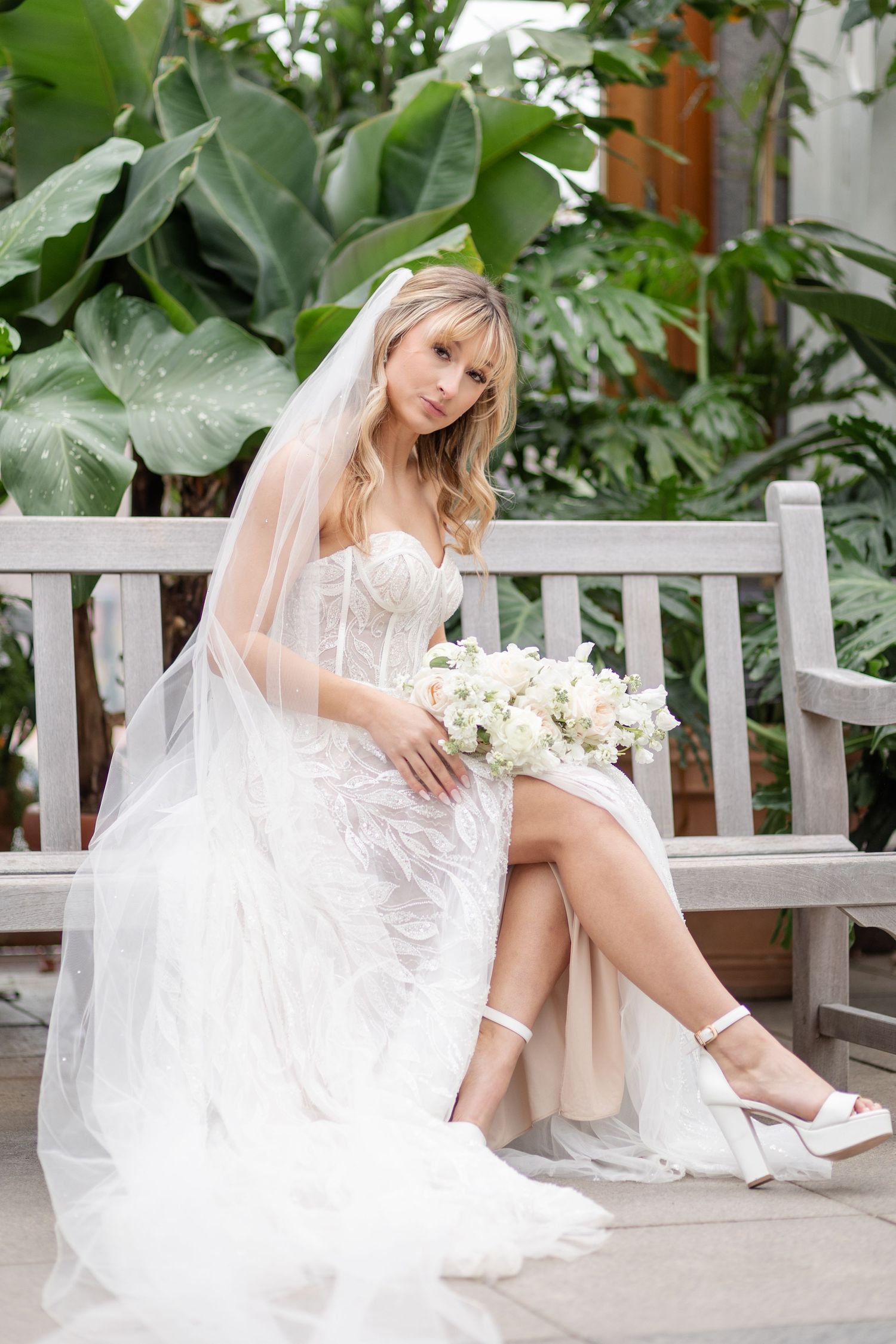 Bride in white lace gown sits on bench with veil and white floral bouquet.
