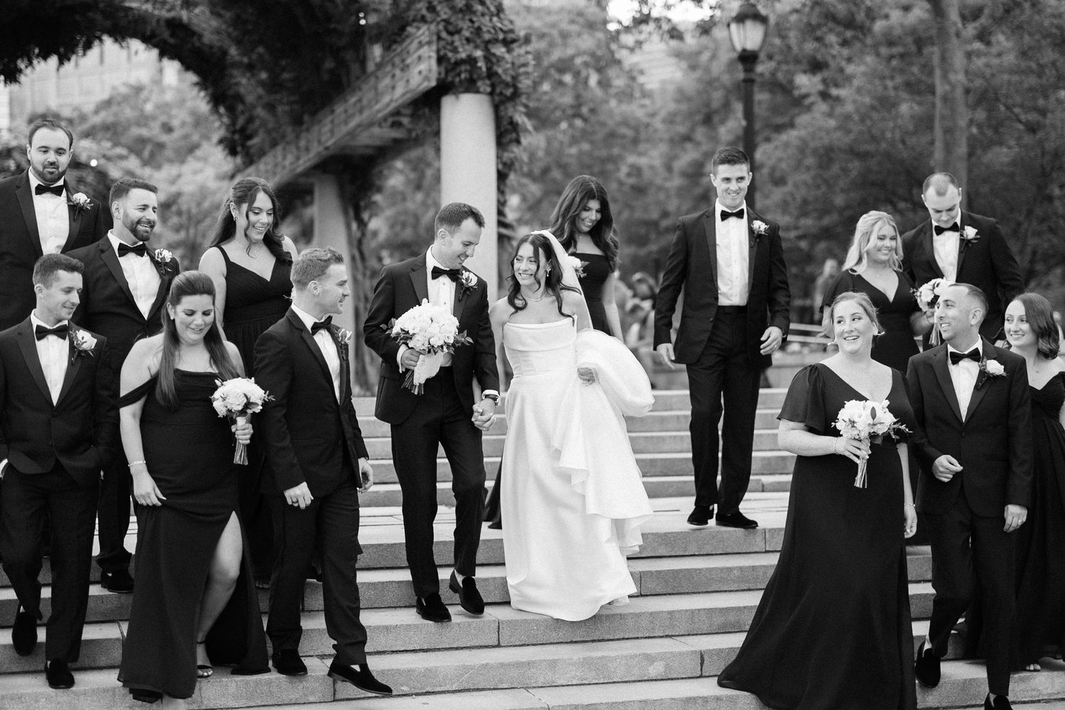 Black and white photo of a wedding party descending outdoor stone steps, bride and groom centered among elegantly dressed guests.