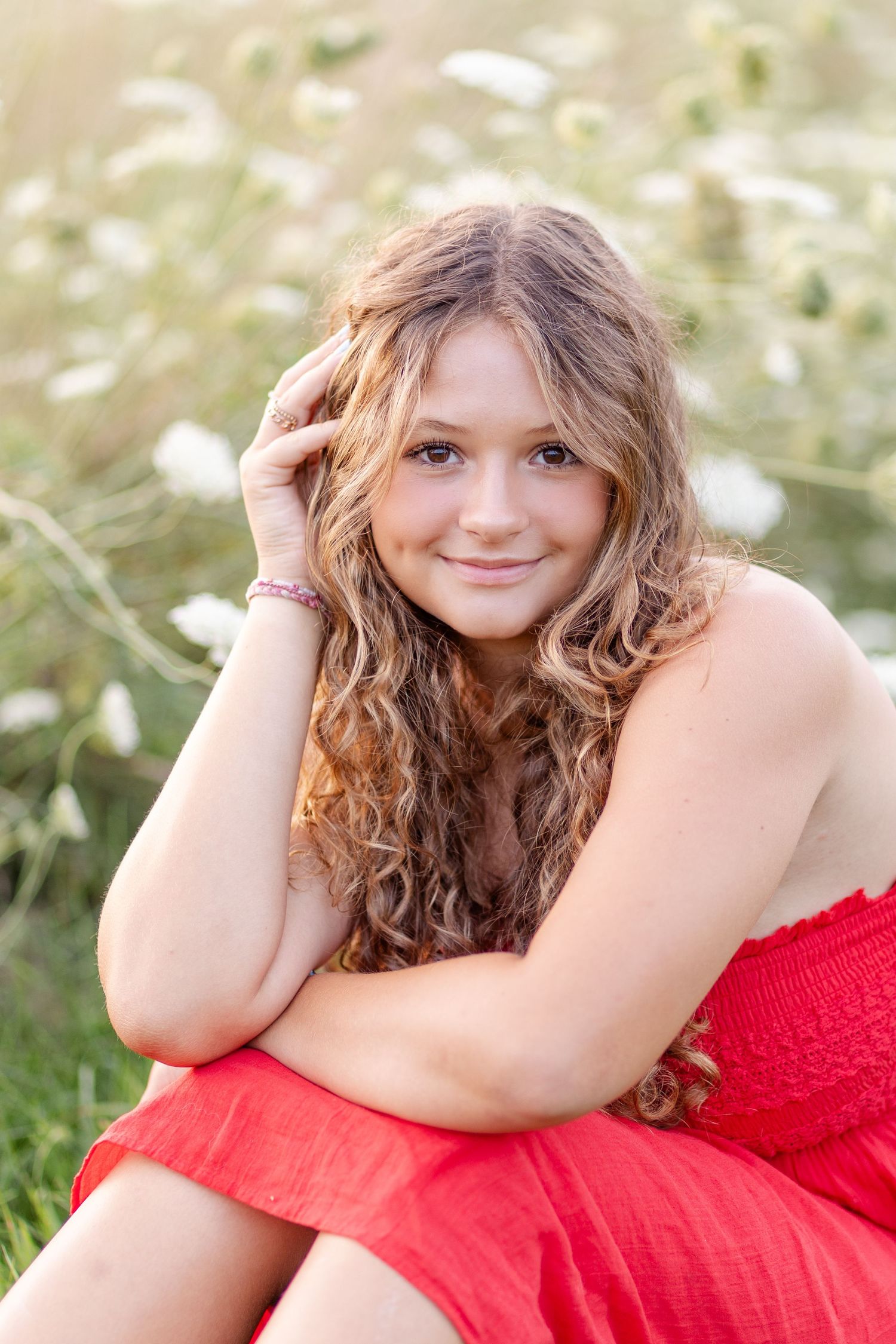 Curly-haired girl in red dress sitting in wildflower field, smiling with hand in hair.