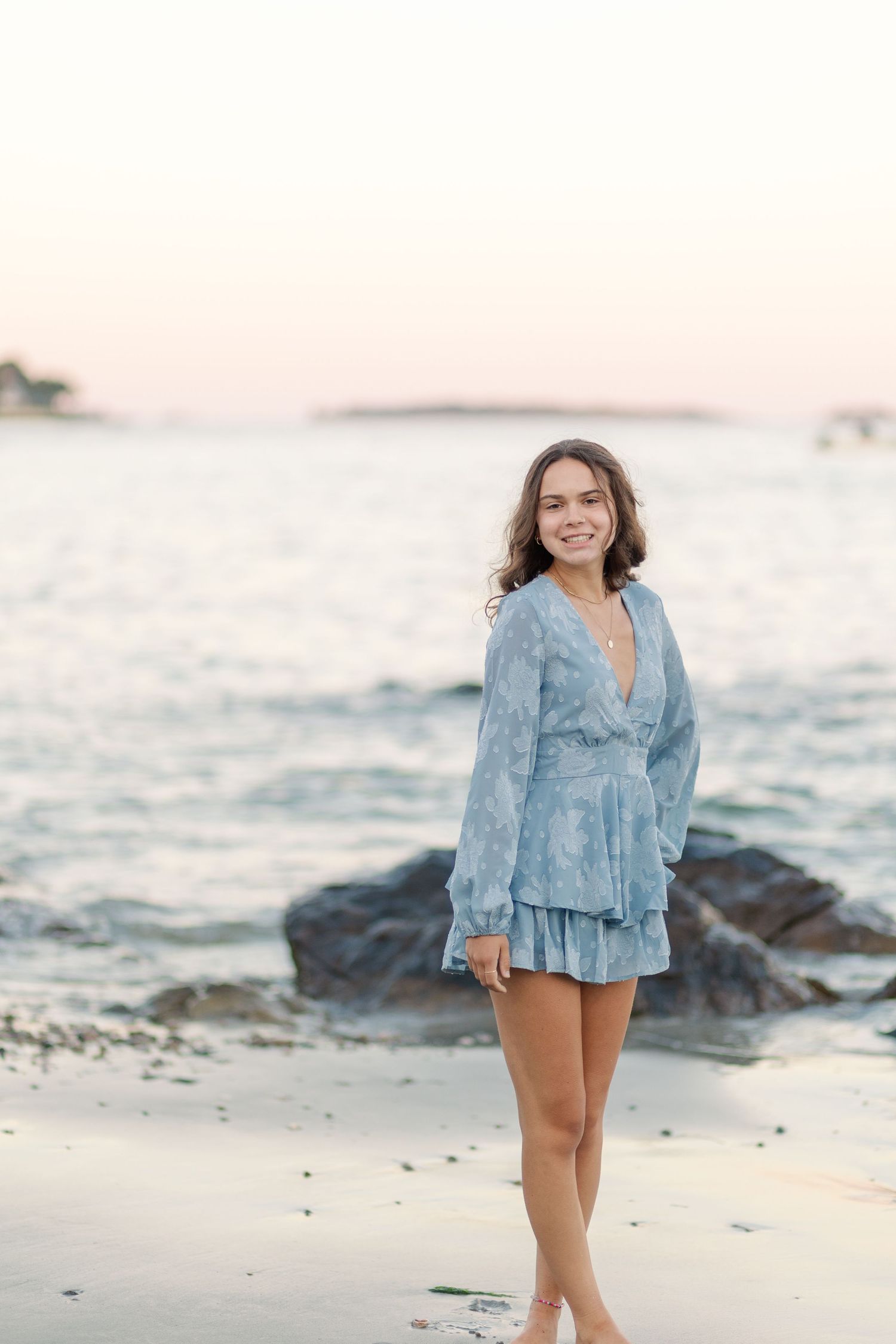 Young woman in light blue long-sleeve mini dress smiling on sandy beach at sunset.