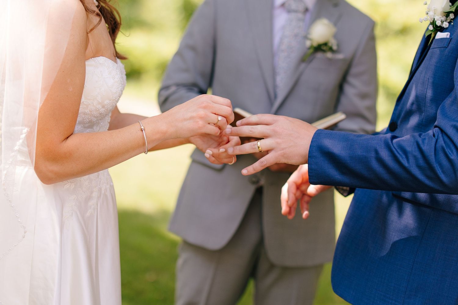 Bride placing wedding ring on groom's finger during outdoor ceremony with officiant in background.