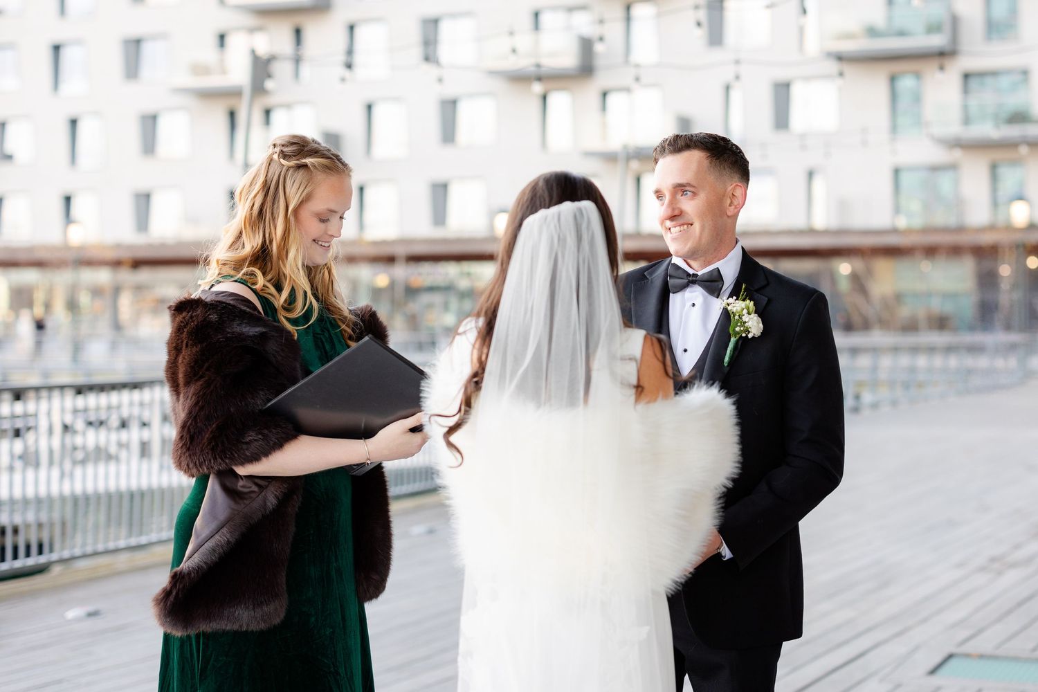 Outdoor winter wedding ceremony with bride in white veil, groom in tuxedo, and officiant in green dress holding book.