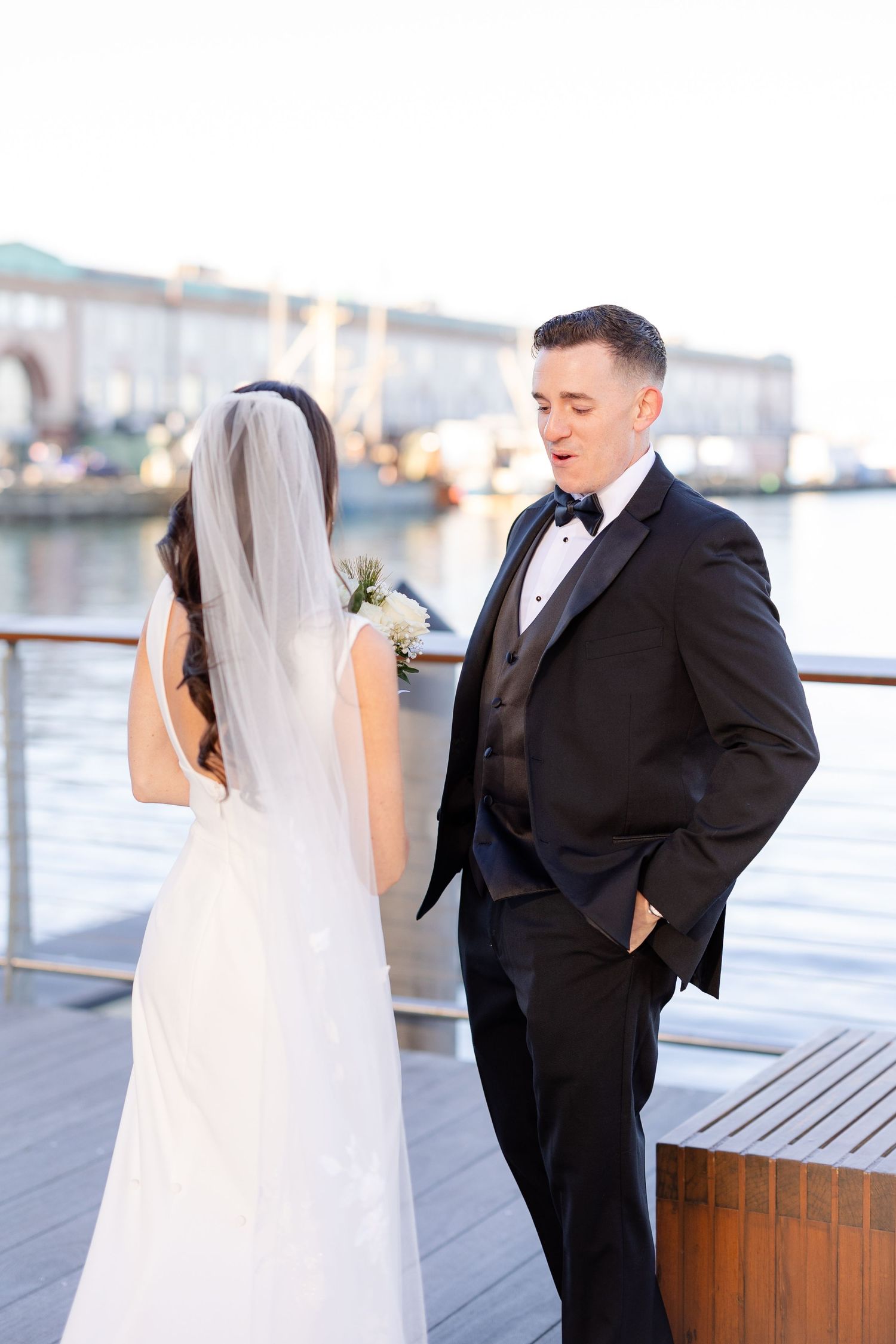 Groom in black tuxedo turns to see bride during first look moment on sunny waterfront dock.