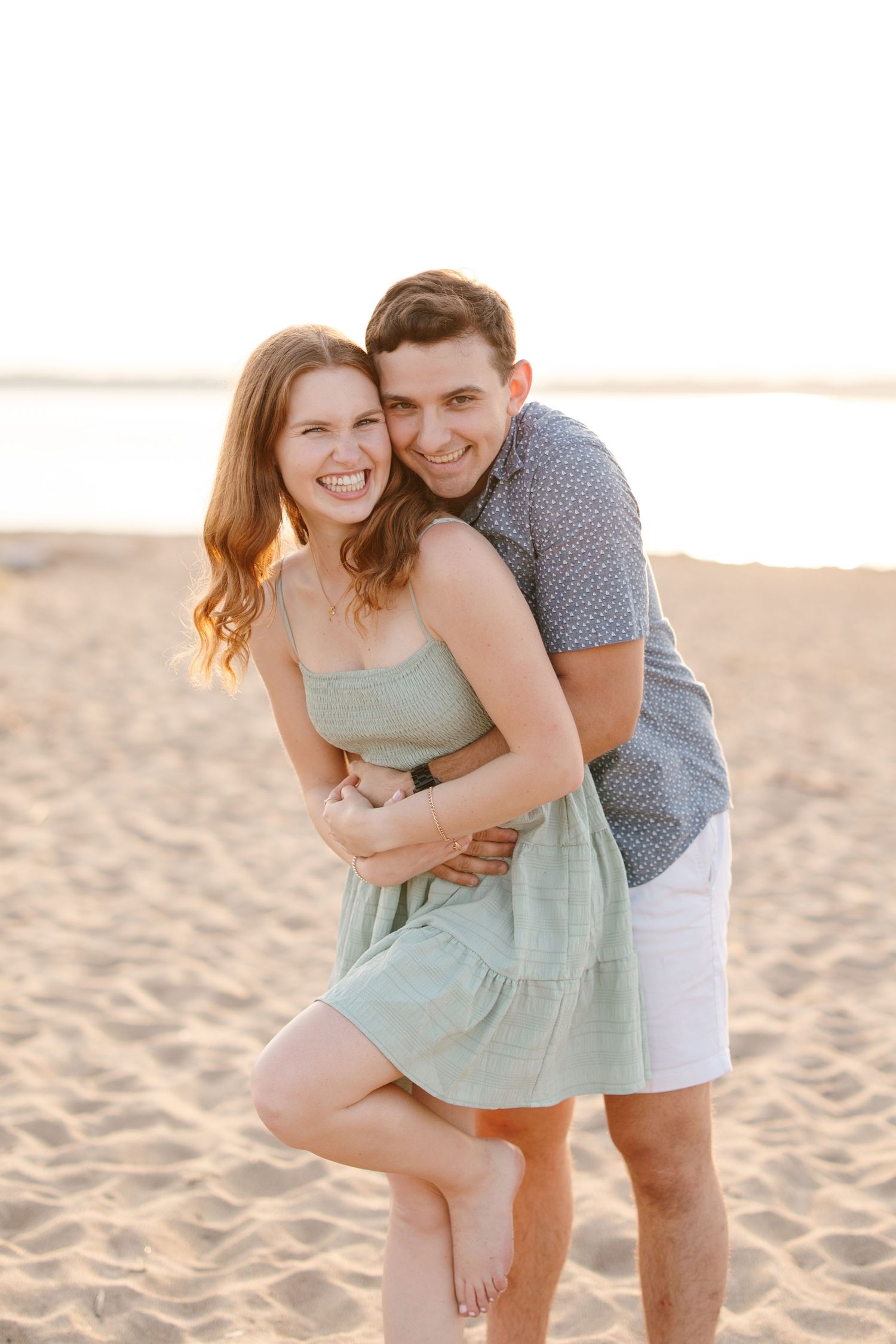 Happy couple embracing on a sunny beach at golden hour, woman in mint green dress laughing.