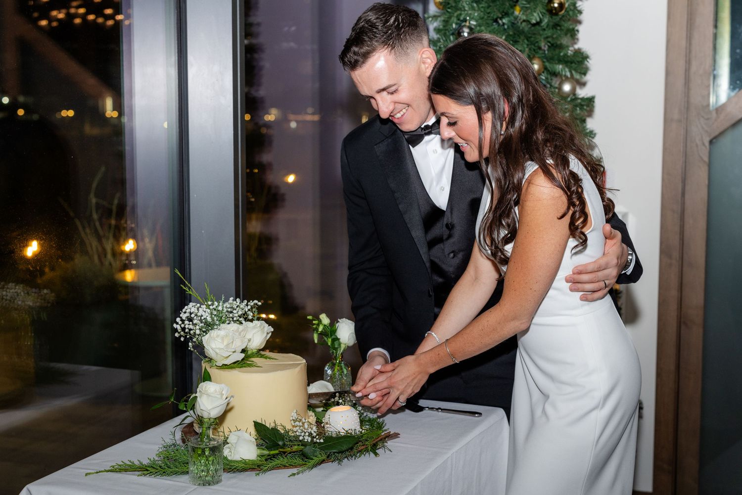 Bride and groom cutting elegant white wedding cake decorated with fresh flowers.