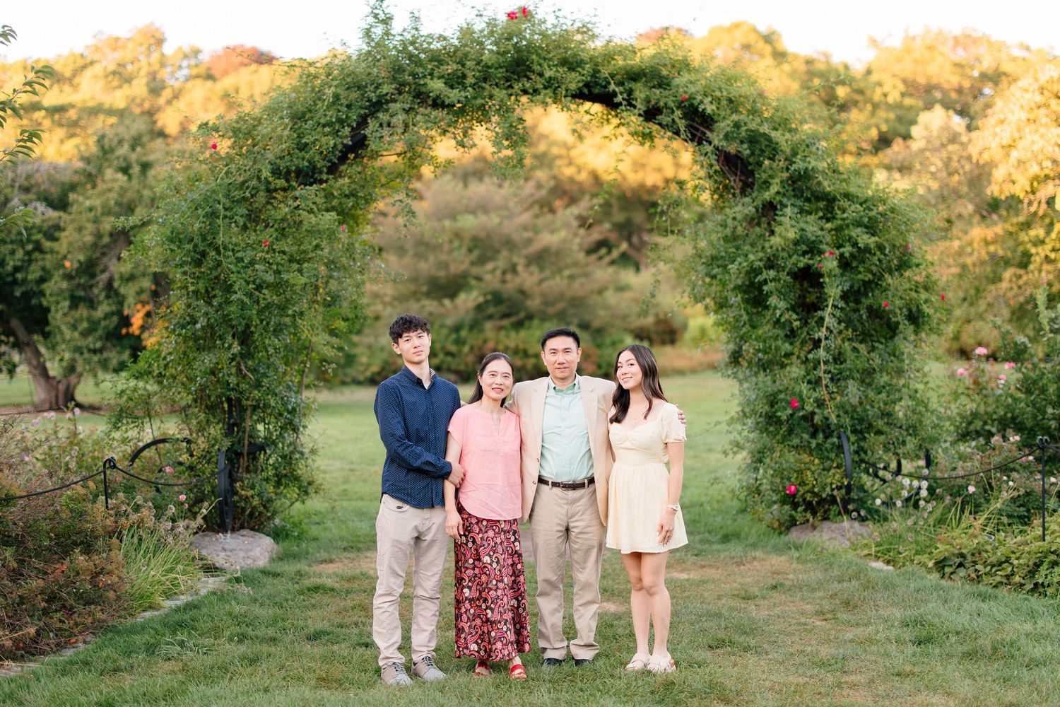 A family of four smiles together beneath a lush green garden arch during an outdoor fall family portrait session.