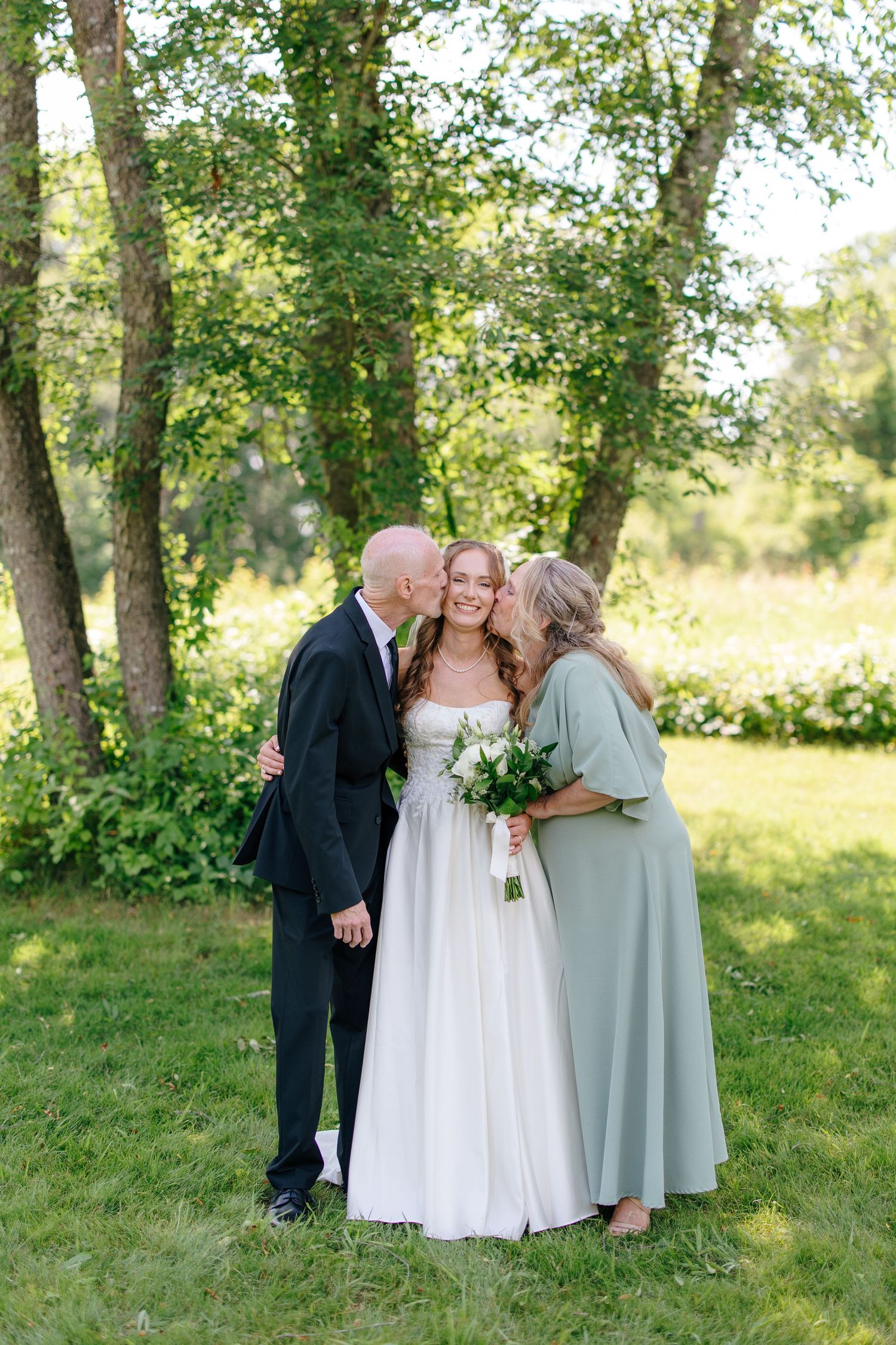 Bride receiving kisses on cheeks from both parents in a lush green outdoor garden setting.