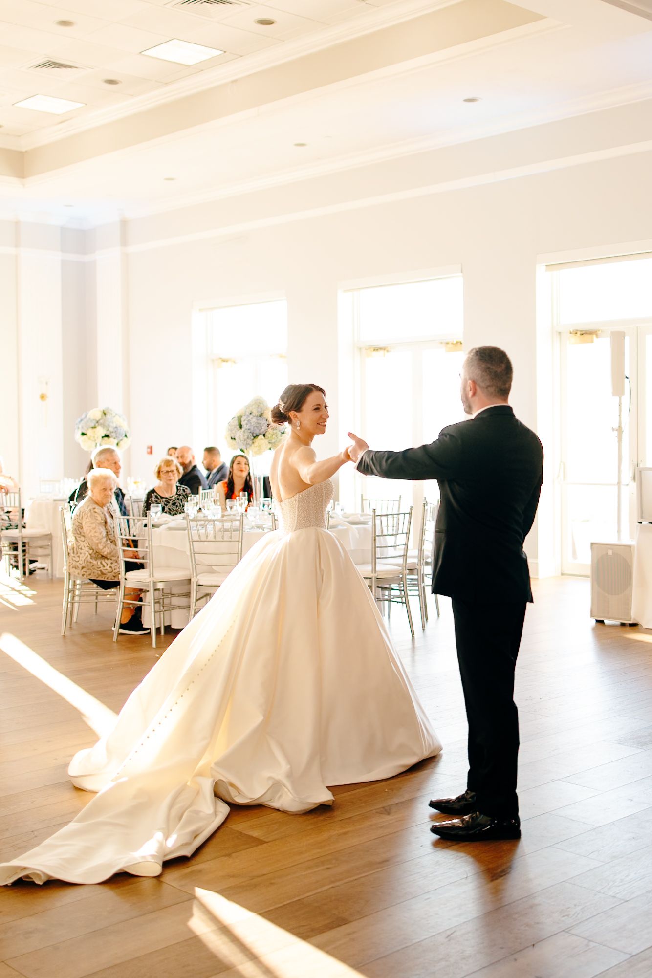 Bride in ballgown and groom sharing first dance in sunlit elegant white wedding reception ballroom.