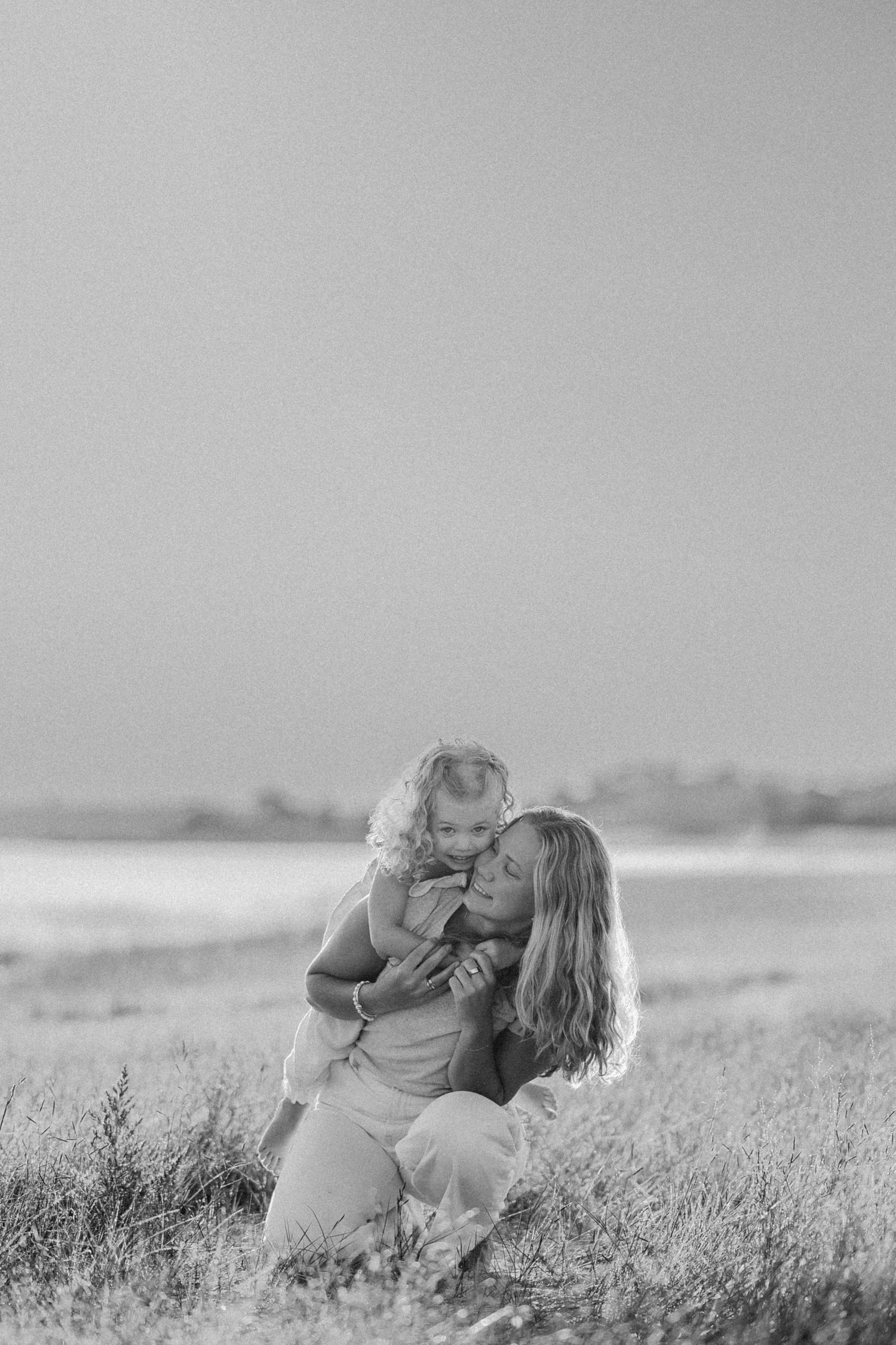Black and white photo of a mother kneeling in a field, lovingly holding her curly-haired toddler close.