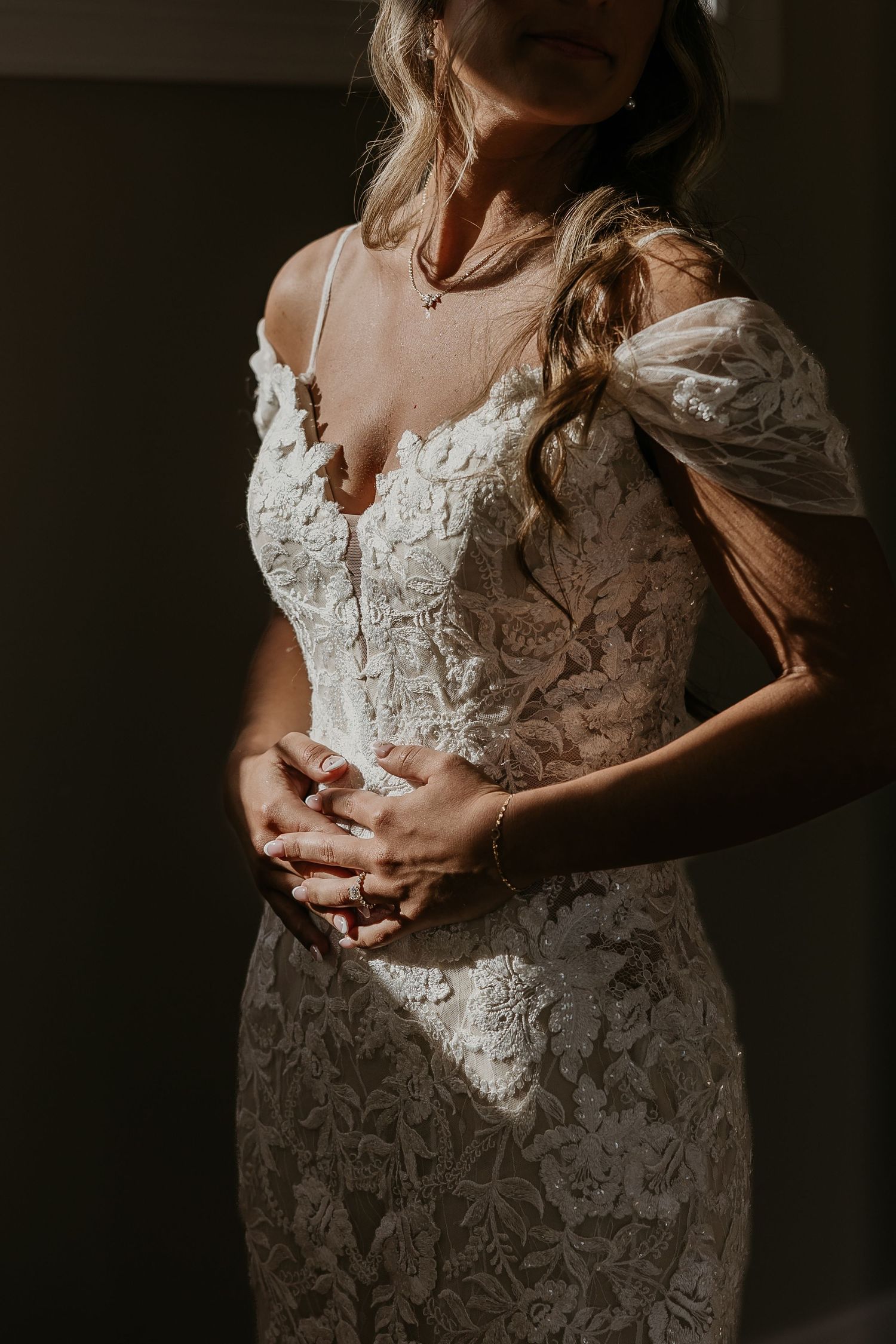Bride in fitted lace wedding gown with off-shoulder sleeves clasping hands in soft light.