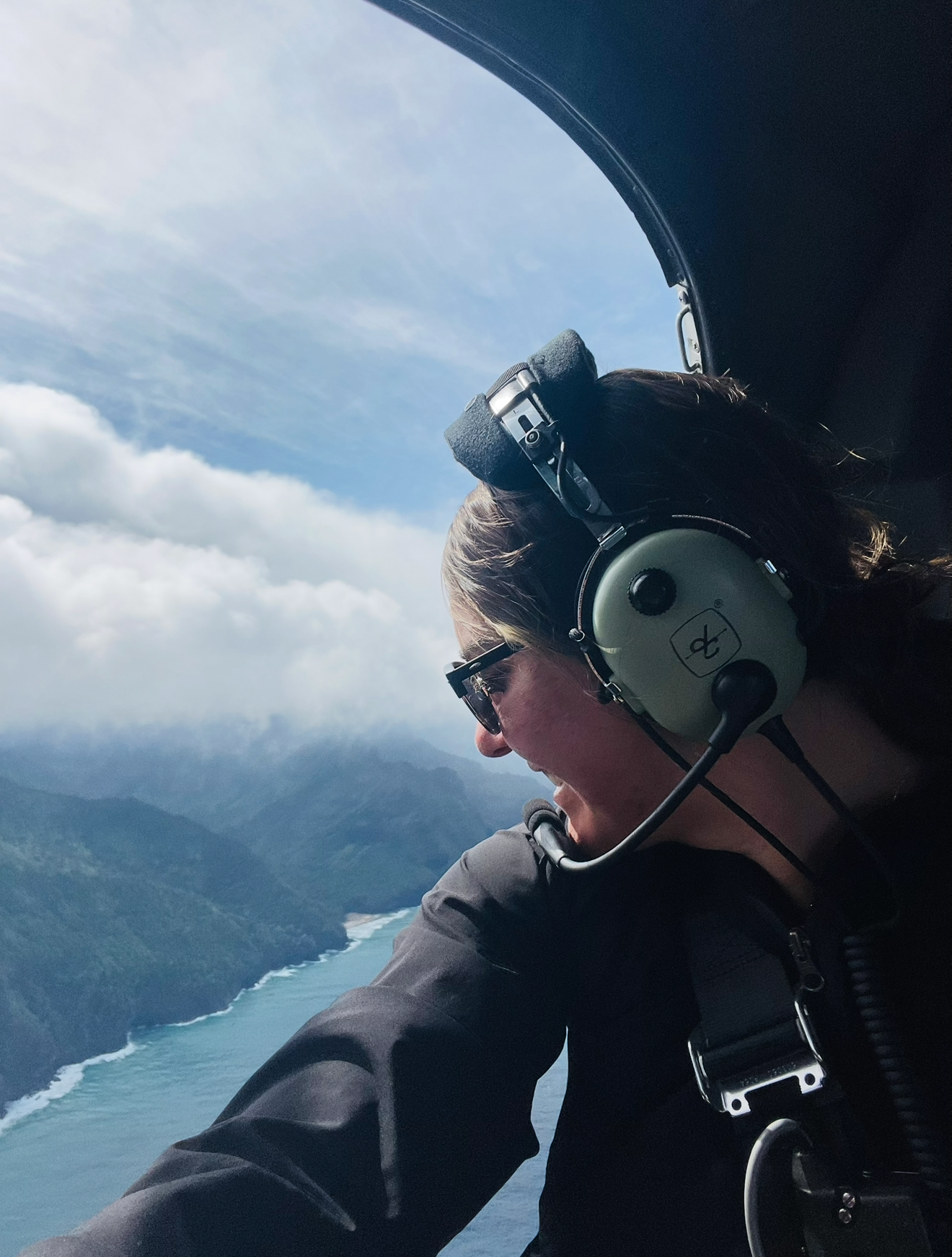 Woman in headset gazes out helicopter window at stunning coastal cliffs and turquoise ocean below.