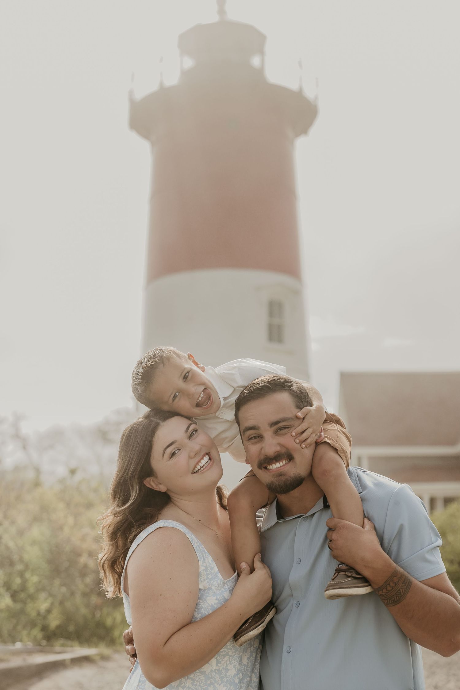Family of three smiling in front of a red and white lighthouse on a sunny day.
