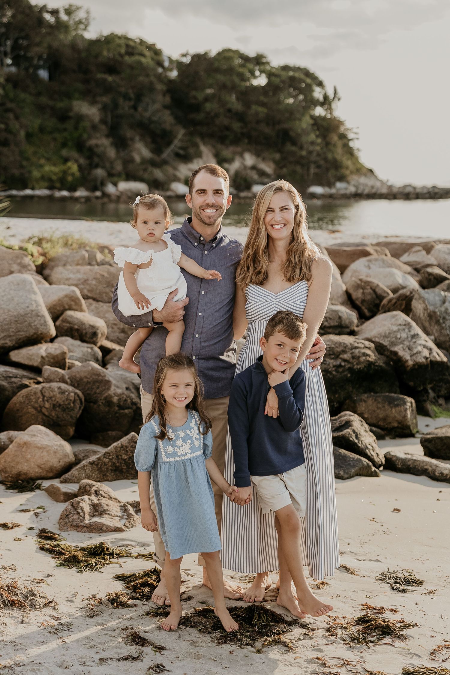 Family of five posing on a rocky beach at sunset, parents with three young children smiling in casual coastal attire.