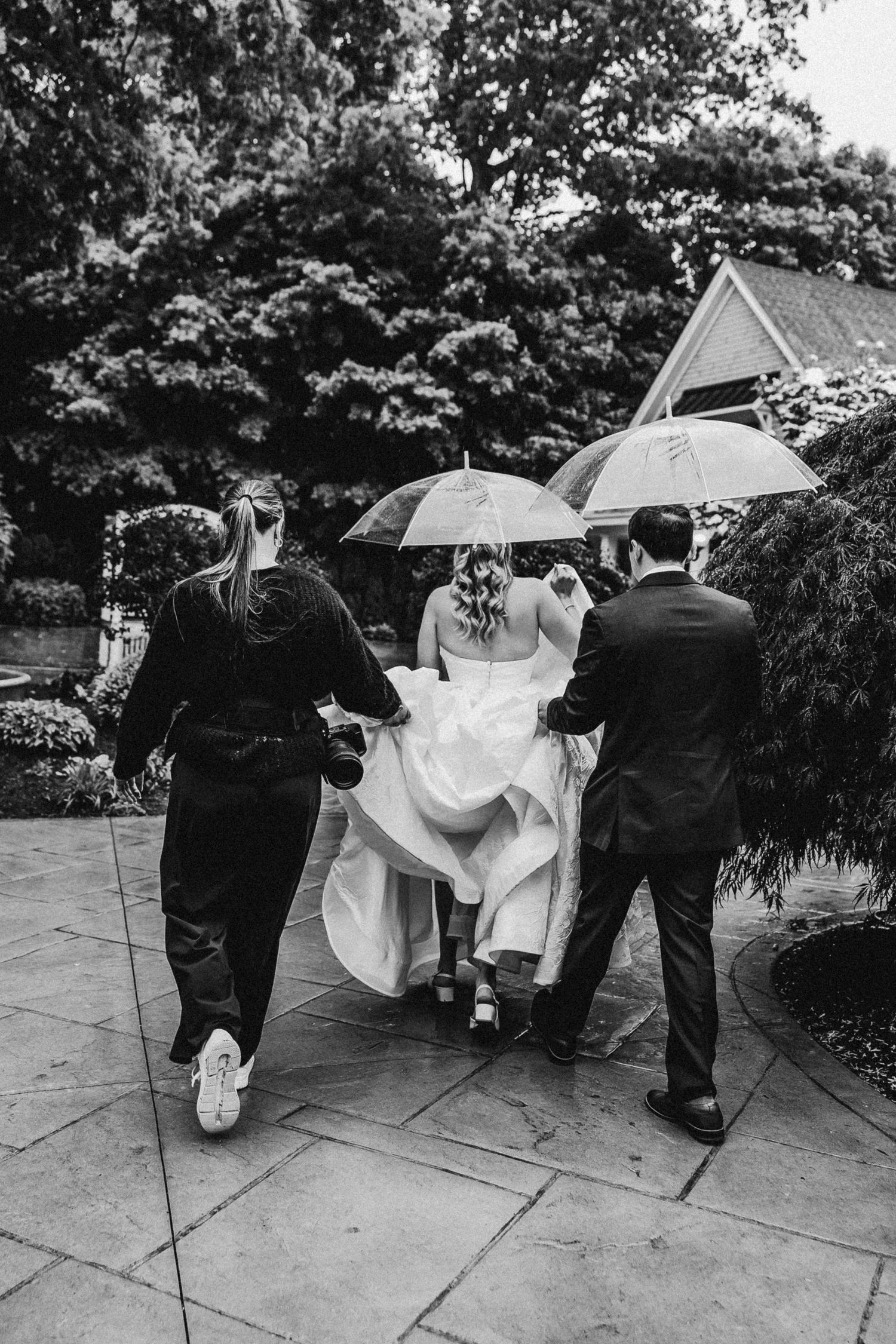 Black and white photo of a bride in a white gown walking with attendants holding umbrellas in a garden.
