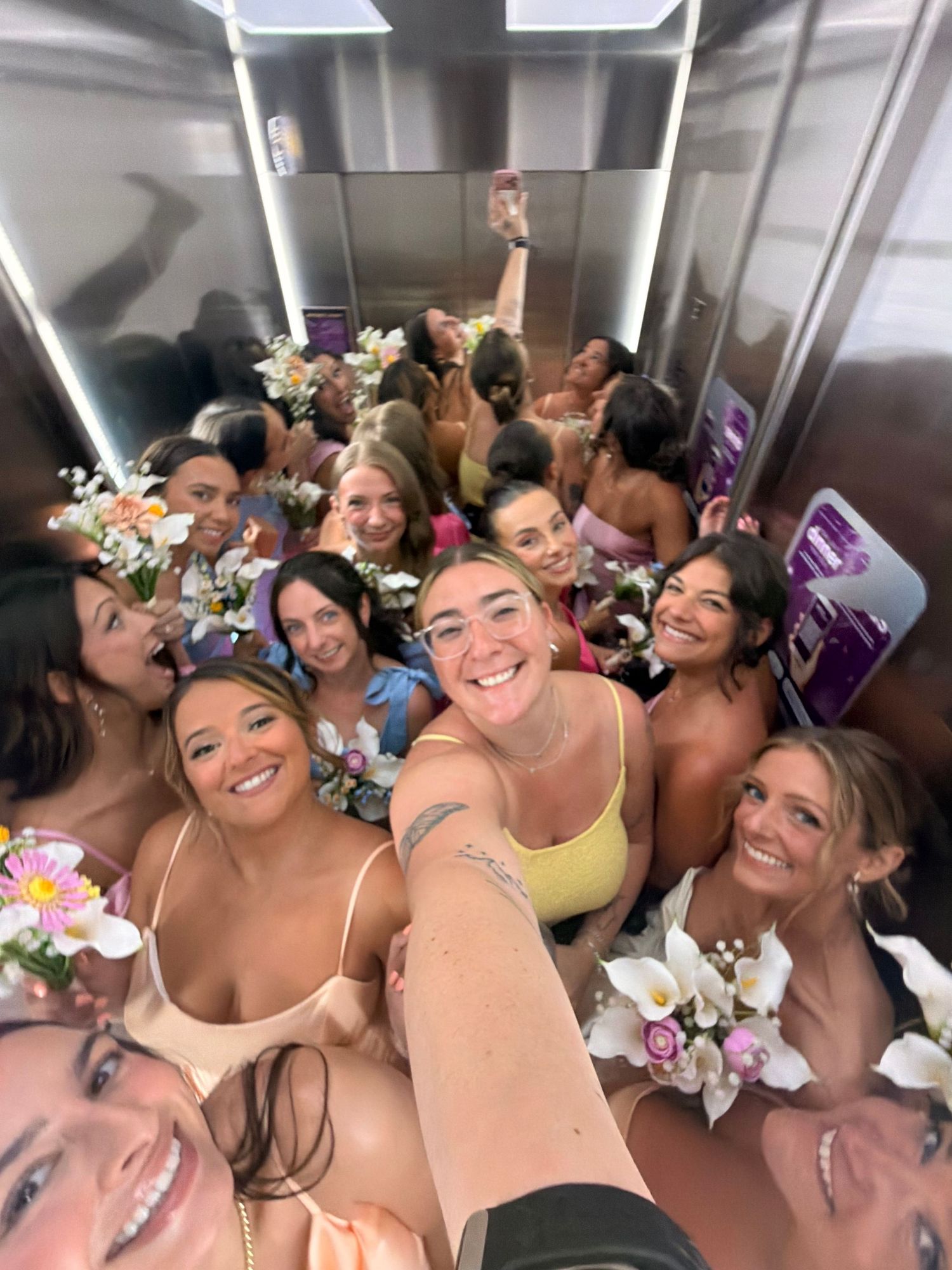 Large group of women taking a selfie in an elevator, wearing swimsuits and floral accessories.