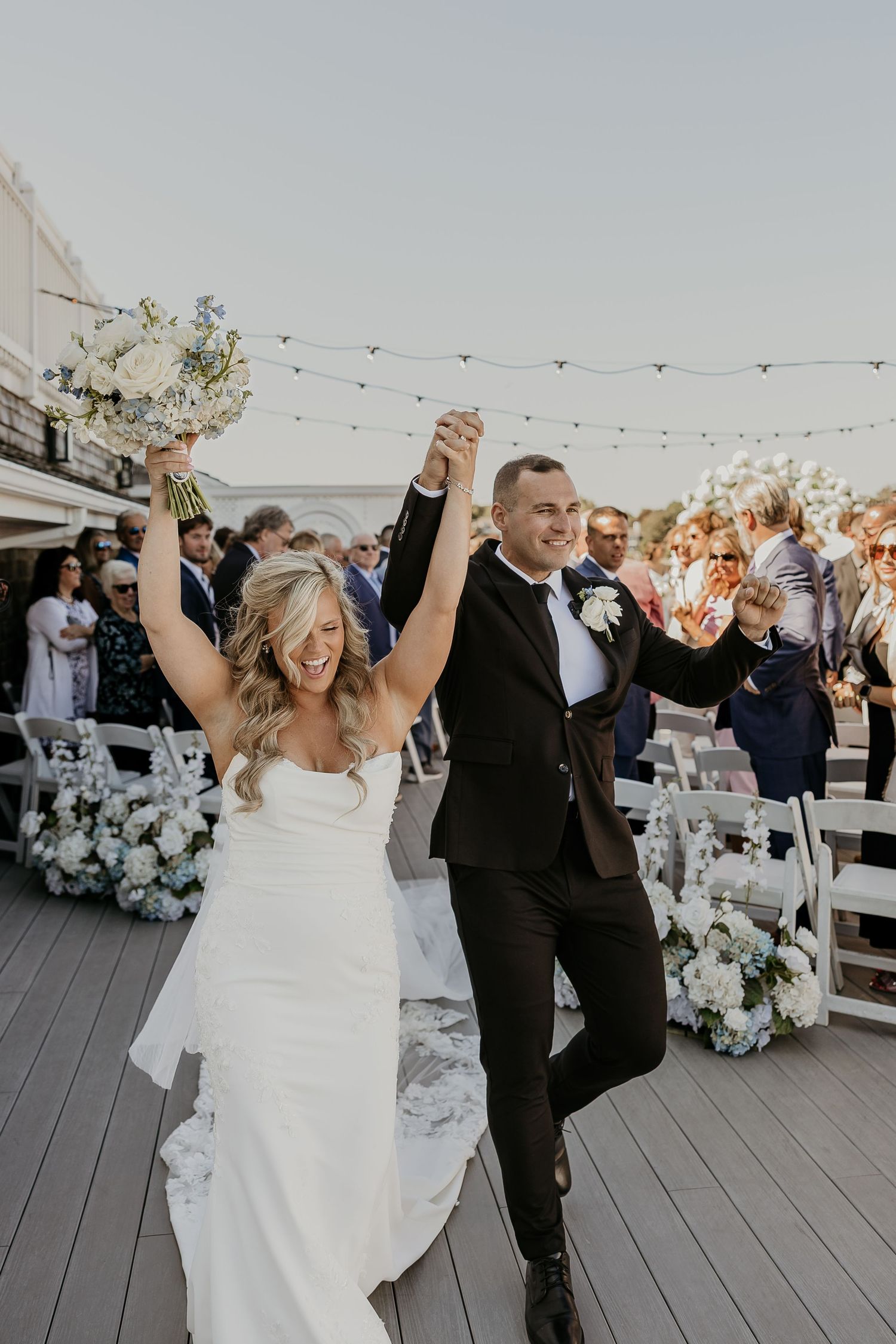 Joyful newlywed couple raising hands in celebration after outdoor rooftop wedding ceremony with guests seated.
