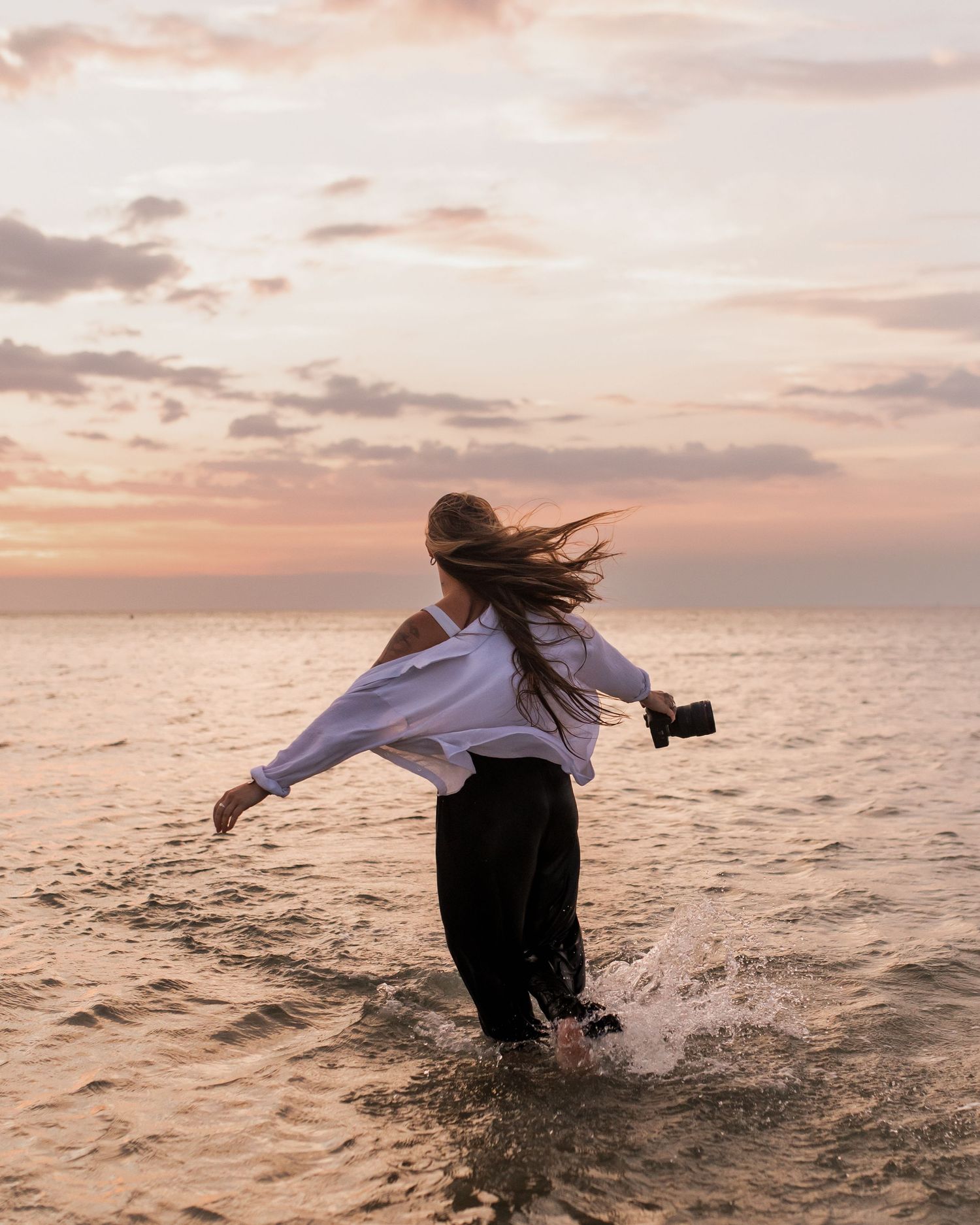Woman wading in ocean at sunset, holding camera, white shirt, black pants, hair blowing in wind.