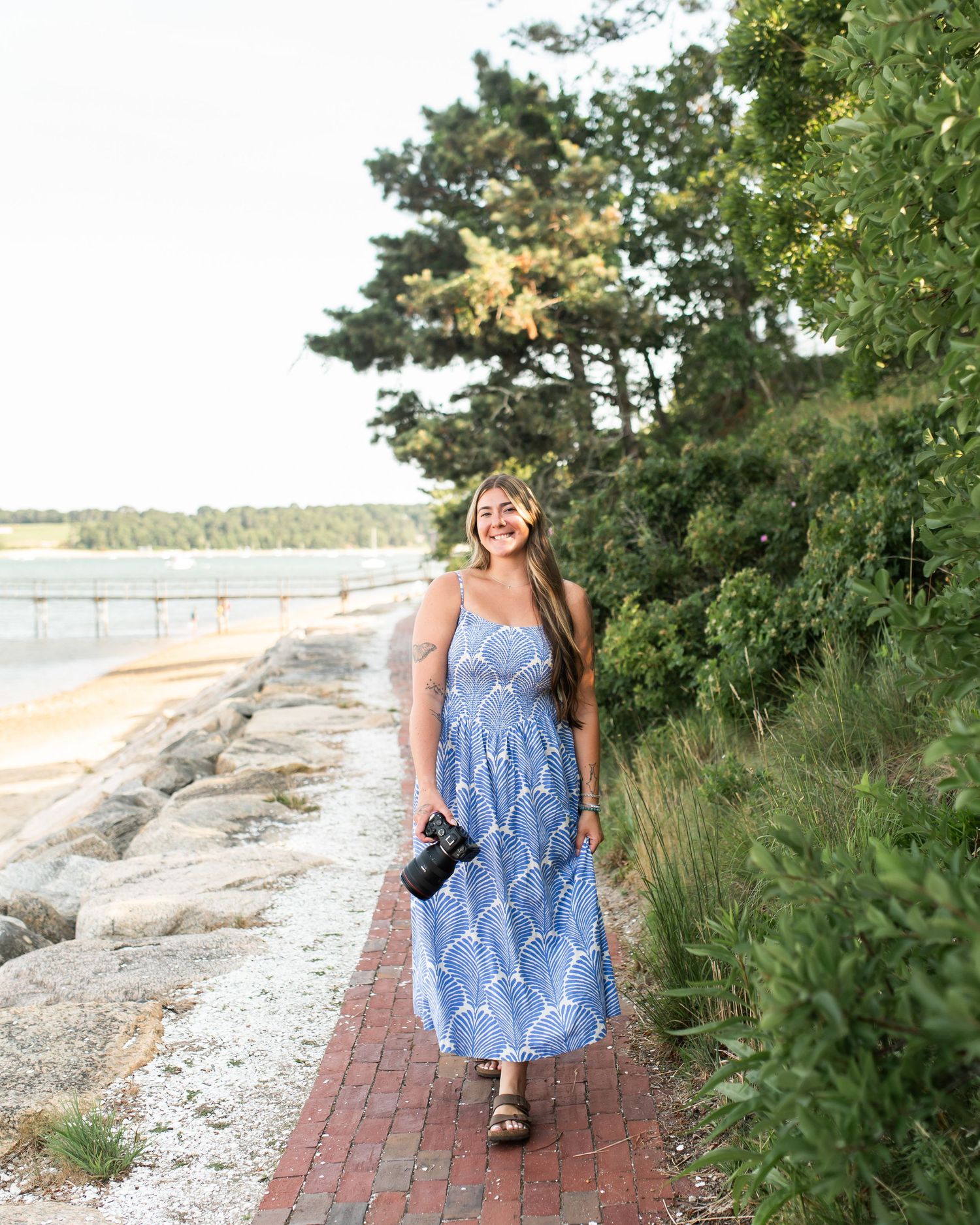Smiling woman in blue floral maxi dress holding camera walks along brick path by rocky shoreline with lush green trees.