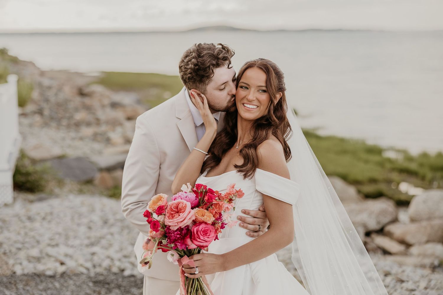 Groom in tan suit kissing bride's cheek as she smiles holding vibrant pink and red bouquet by rocky waterfront.