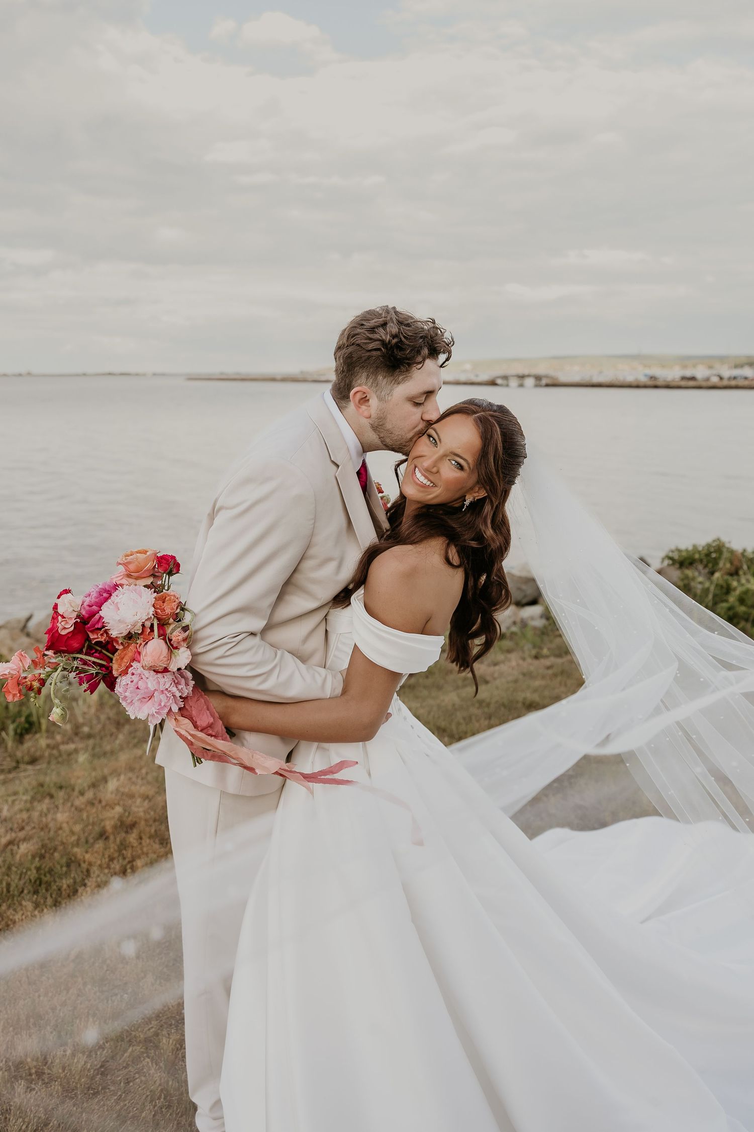 Bride in off-shoulder gown with flowing veil holds pink bouquet as groom in tan suit kisses her cheek by the water.