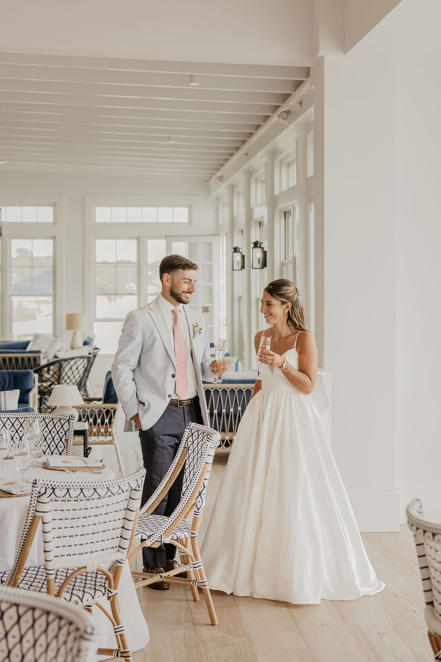 Bride and groom share a joyful moment in an elegant coastal restaurant, she in a white gown, he in a gray suit.