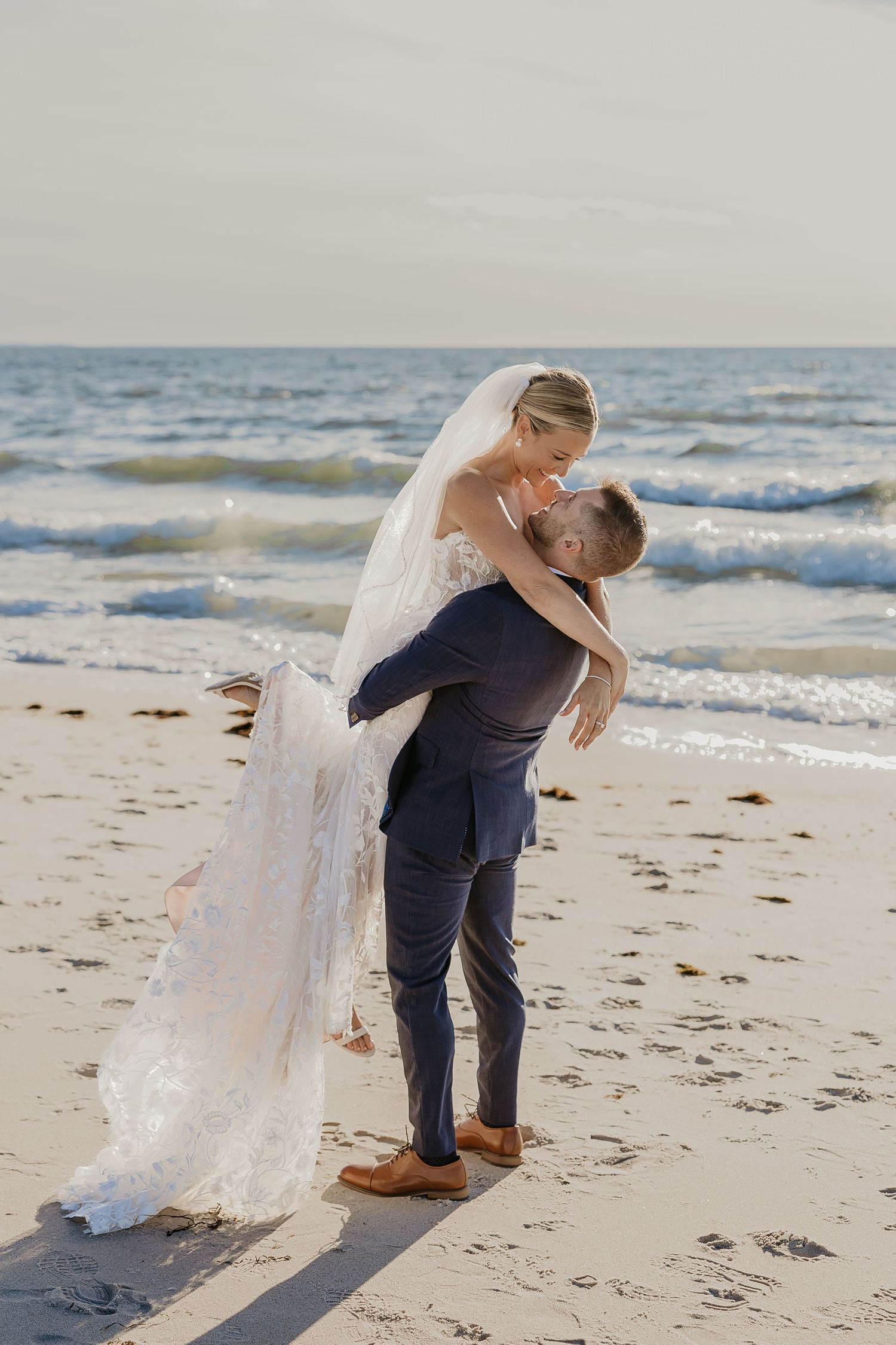 Groom lifting bride in white lace gown and veil on sandy beach with ocean waves, romantic wedding photo.