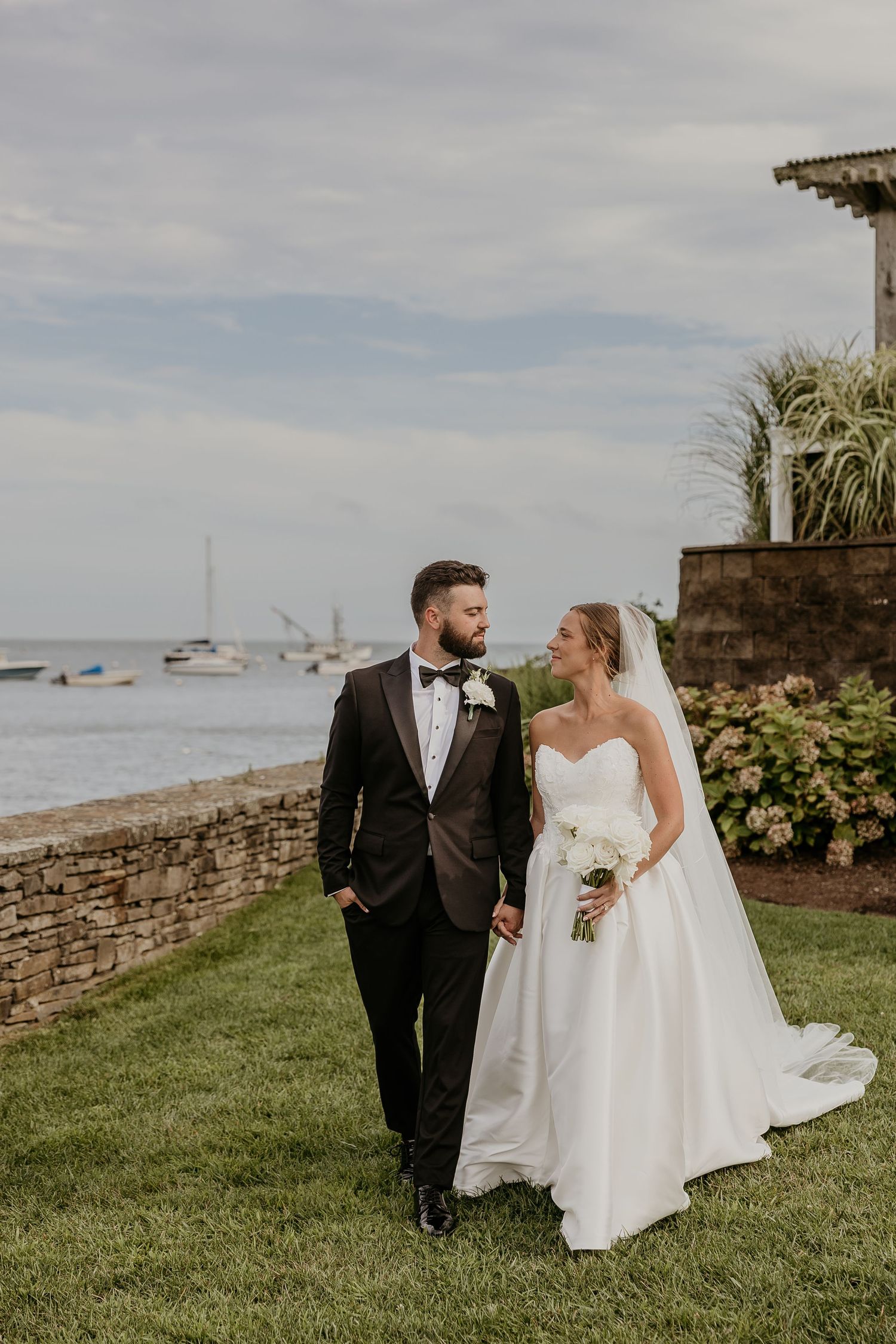 A bride and groom walk hand-in-hand on a waterfront lawn, she in a white gown, he in a dark tuxedo.