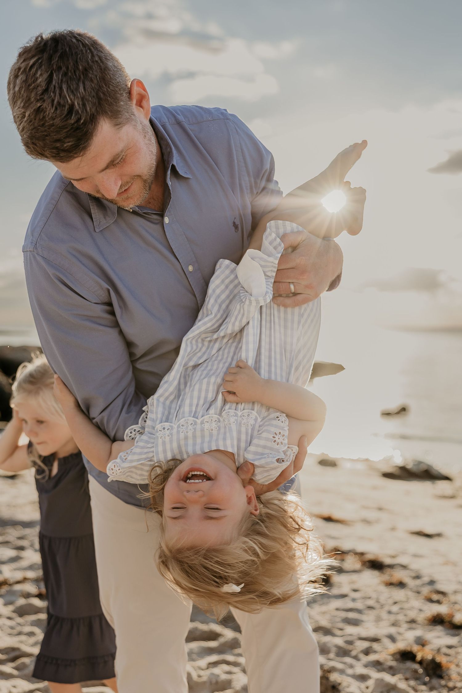Father playfully holds child upside down on sunny beach while another child stands nearby.