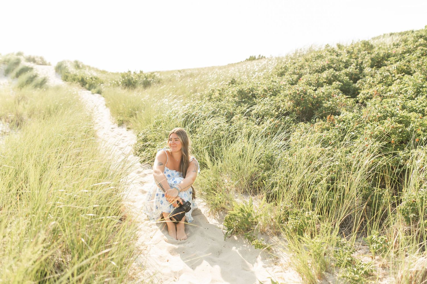 Woman sitting on a sandy coastal path surrounded by tall green dune grass on a bright sunny day.