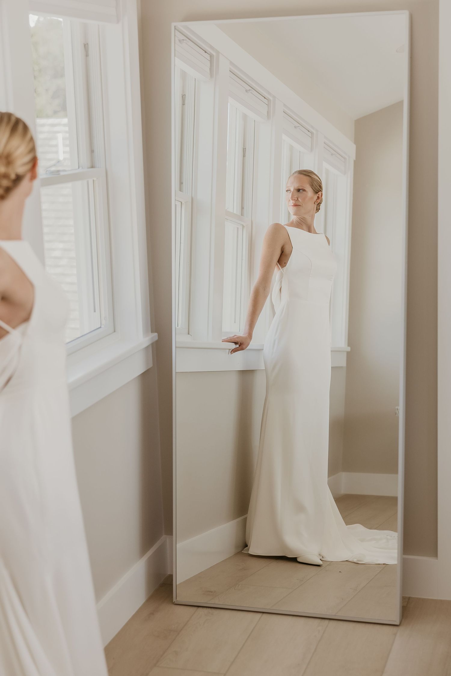 Bride in a minimalist sleeveless white satin gown reflected in a full-length mirror indoors.