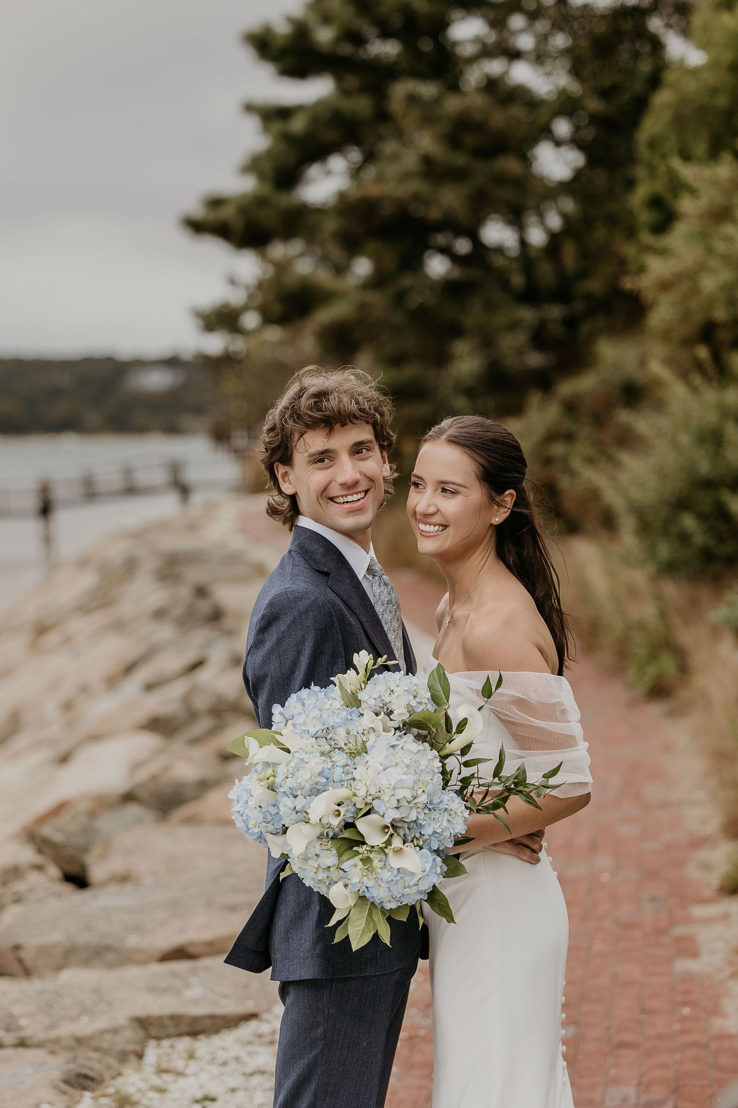 Smiling couple in wedding attire poses by rocky New England shoreline with blue hydrangea bouquet.