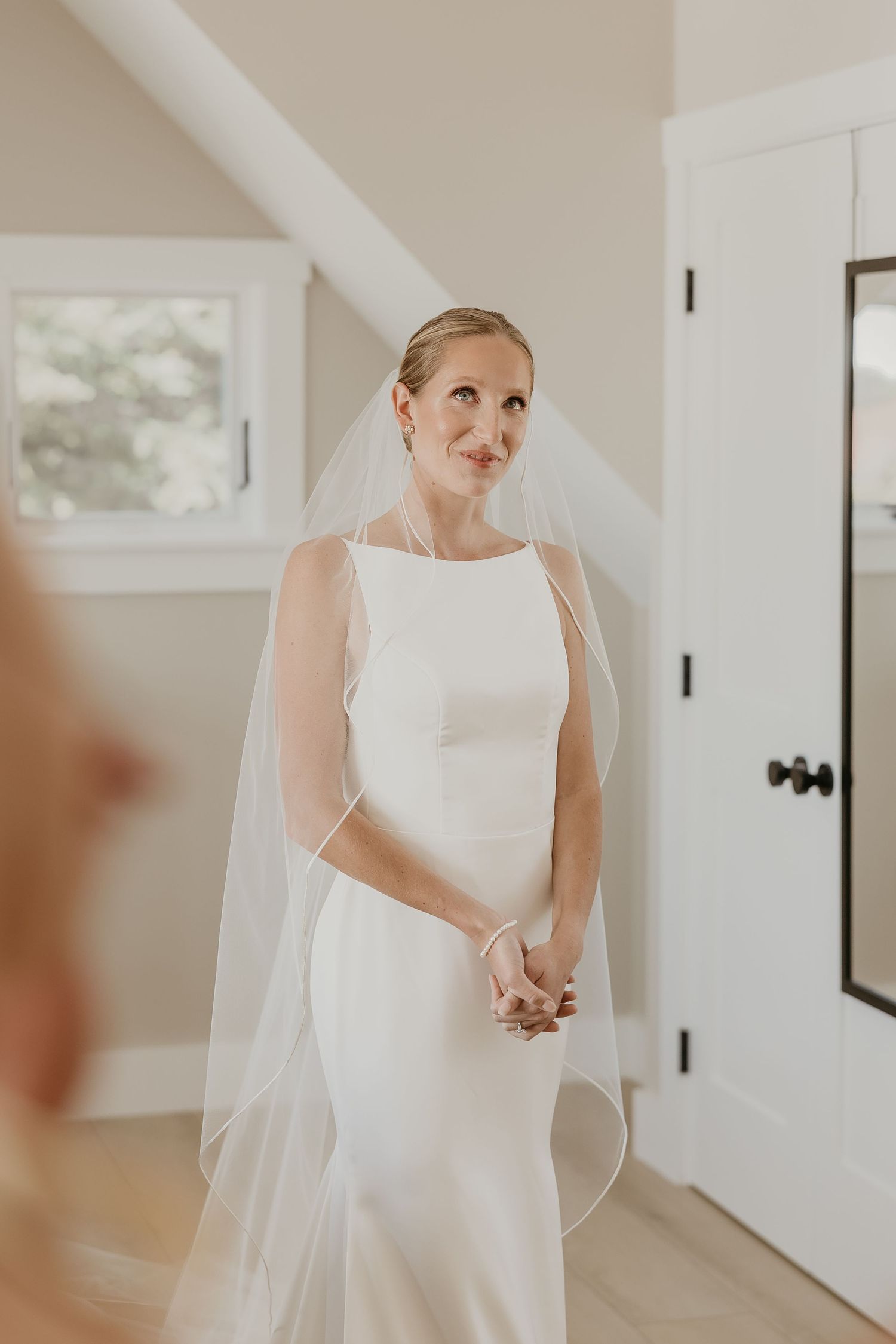 Elegant bride in a simple sleeveless white satin gown with a long veil standing indoors.