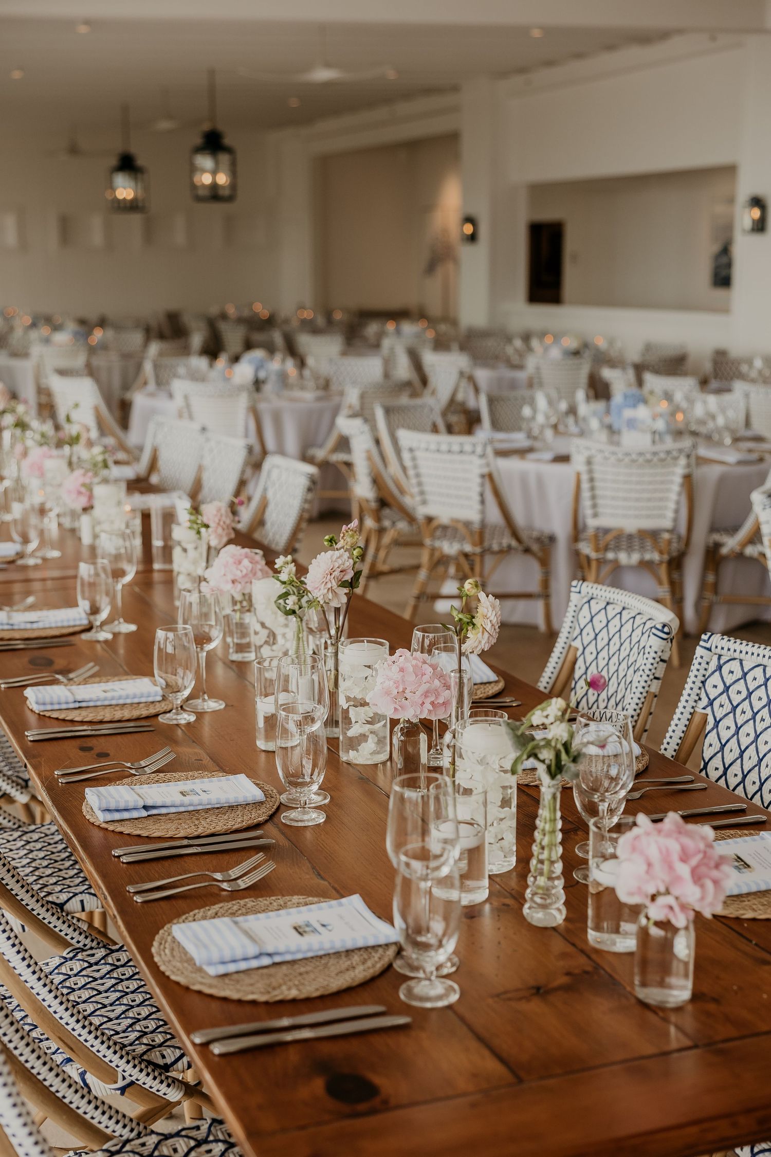 Elegant wedding reception hall with long wooden tables, pink floral centerpieces, crystal glassware, and blue patterned chairs.