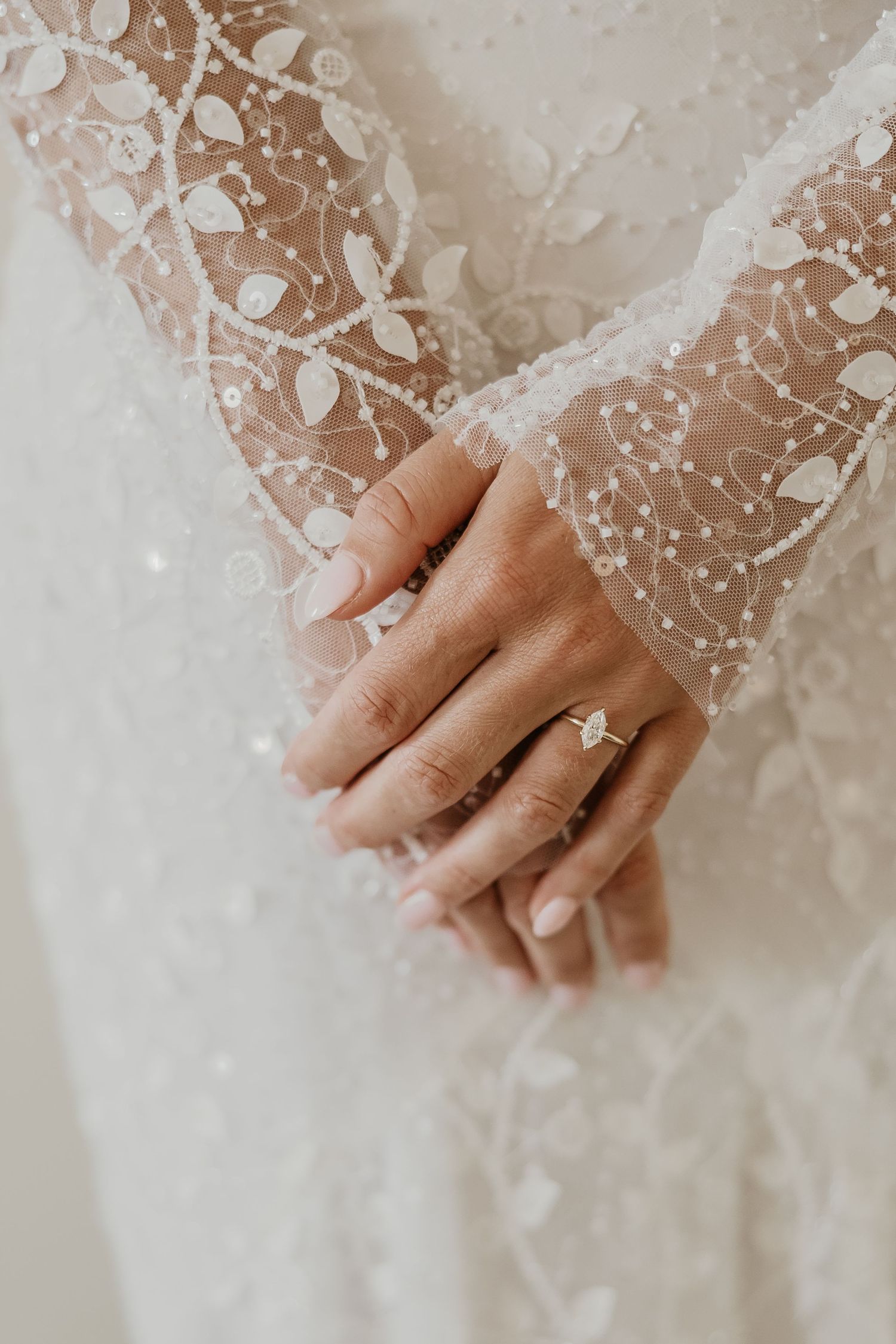 Close-up of a bride's hands wearing a diamond engagement ring over a beaded lace wedding gown.