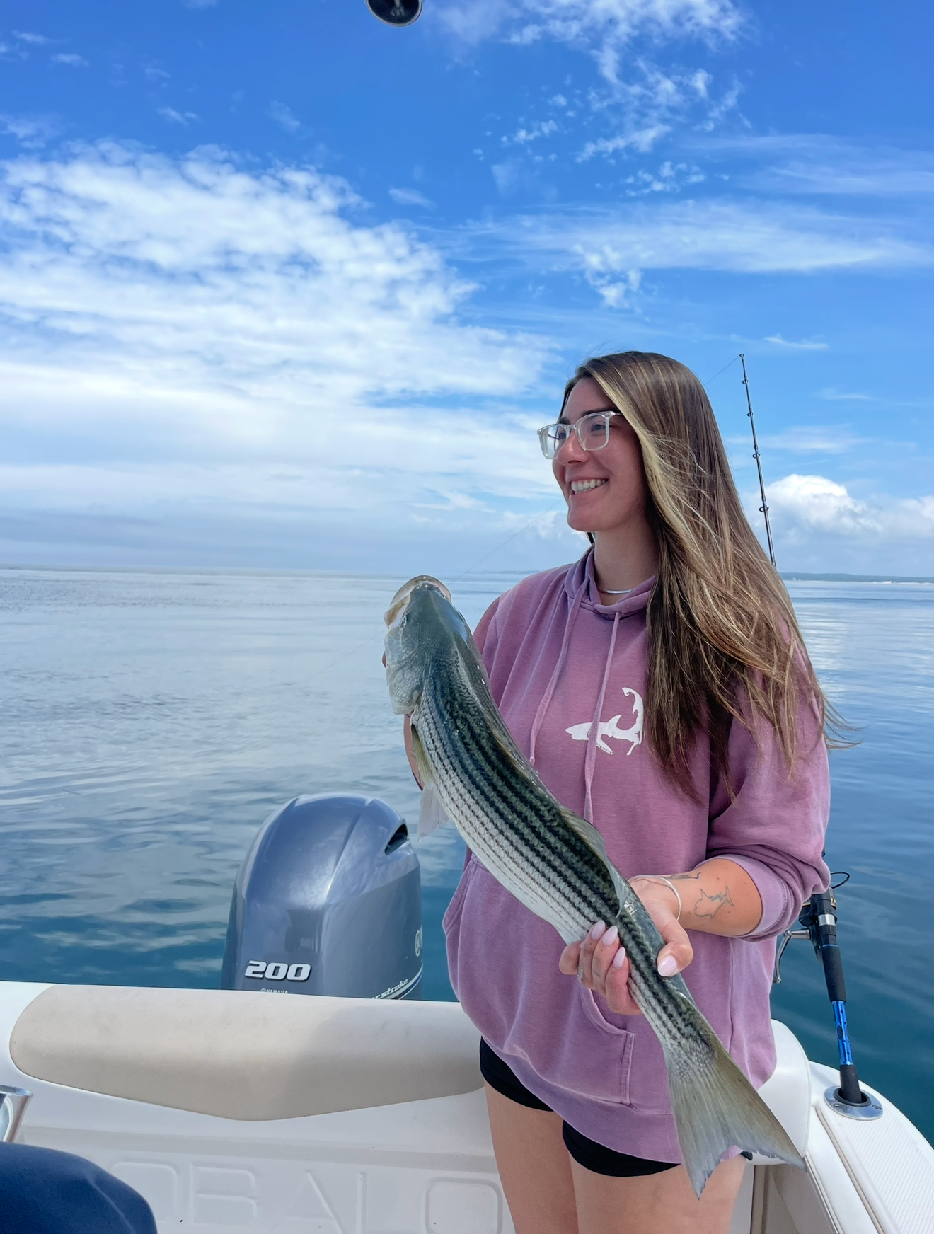 Smiling woman holds large striped bass fish on a boat on calm open ocean waters.