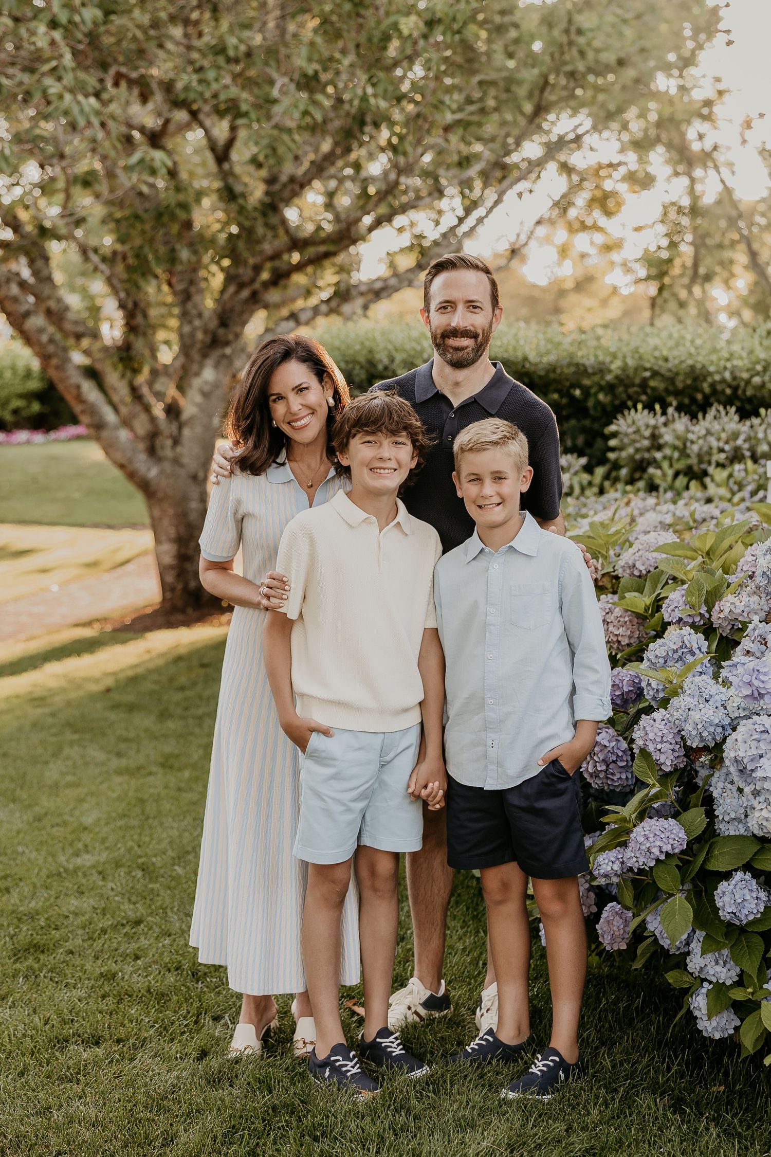 Family of four posing outdoors near blooming hydrangeas and a large tree on a sunny day.