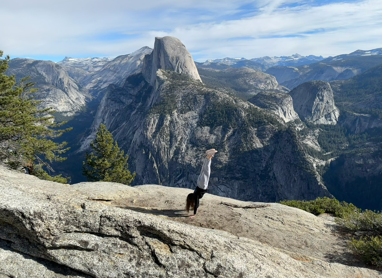 Person poses on rocky overlook at Yosemite with iconic Half Dome mountain peak in the distance.