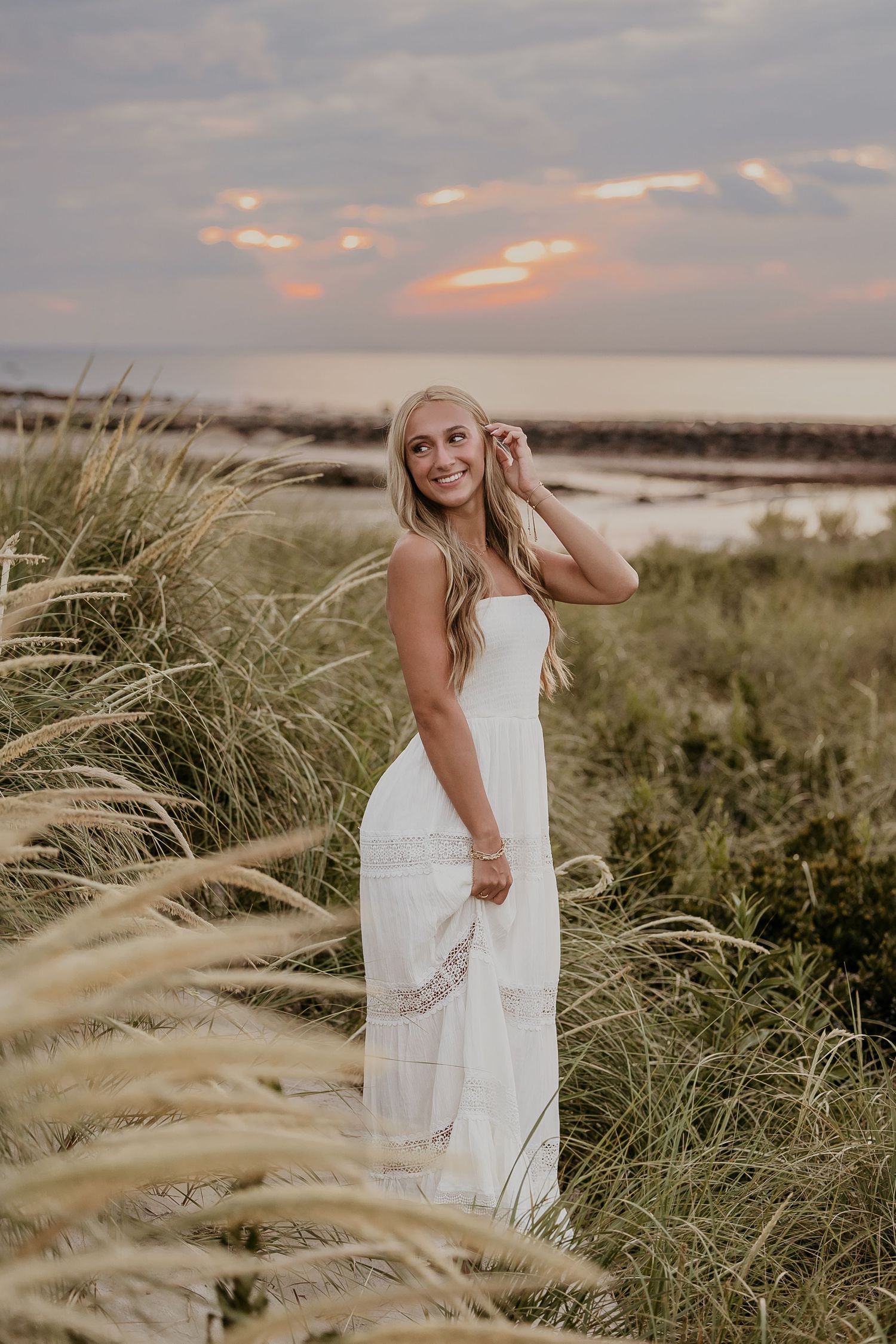 Blonde woman in white maxi dress standing in tall beach grass at sunset near the ocean.