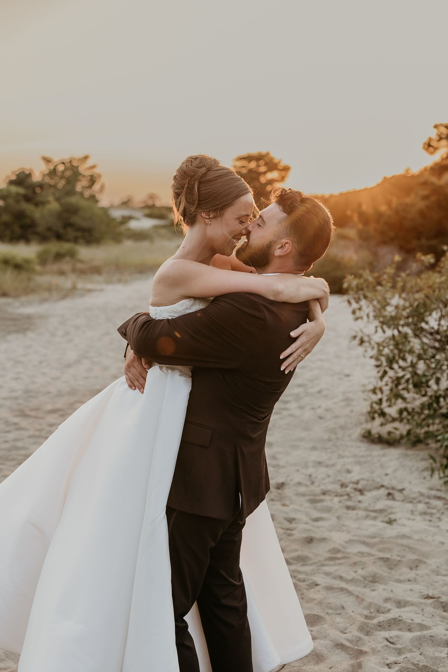 A groom lifts his bride in a white gown at sunset on a sandy beach, sharing a romantic moment.