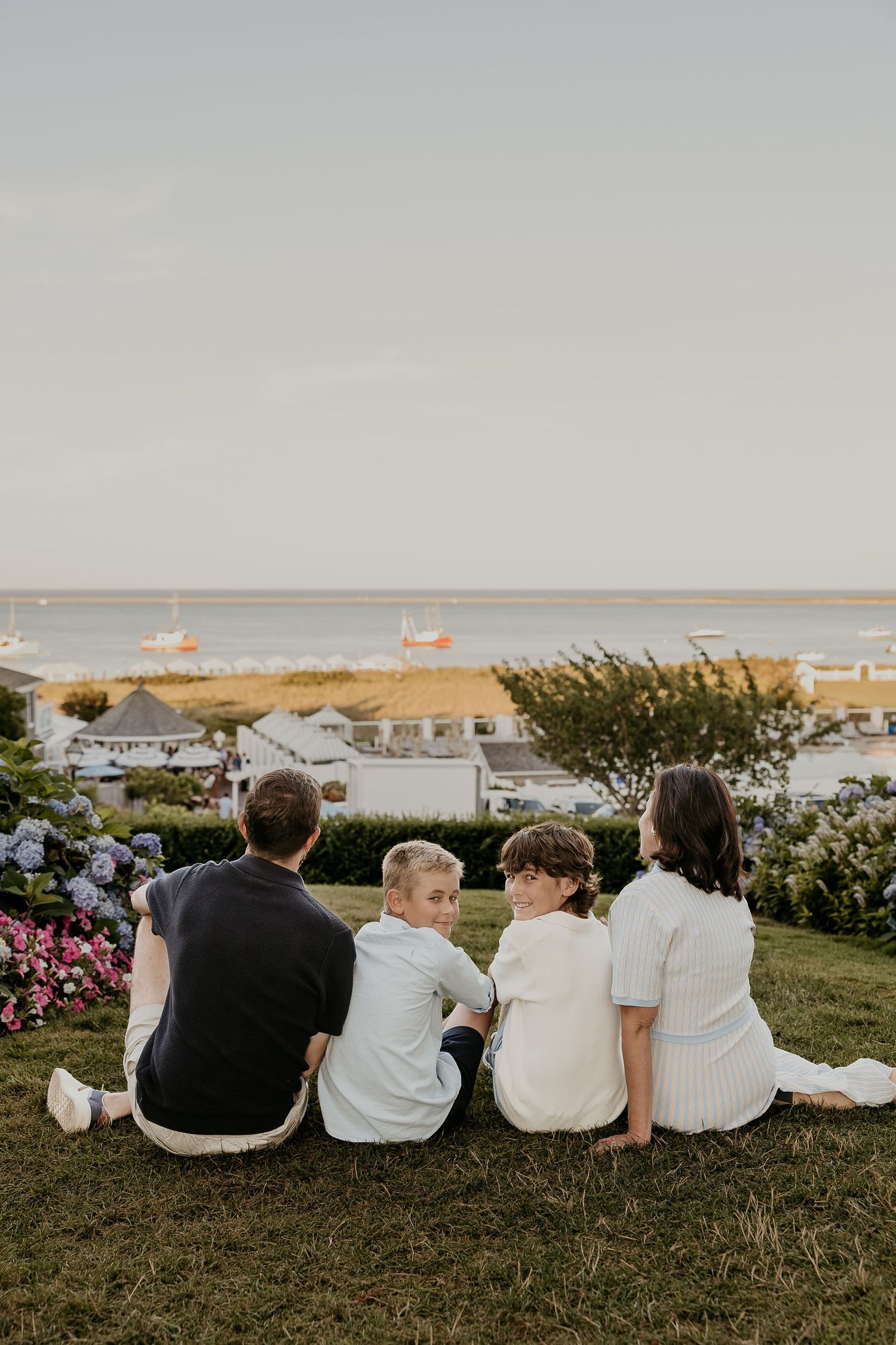Family of four sitting on a grassy hill overlooking a scenic coastal harbor with boats at golden hour.