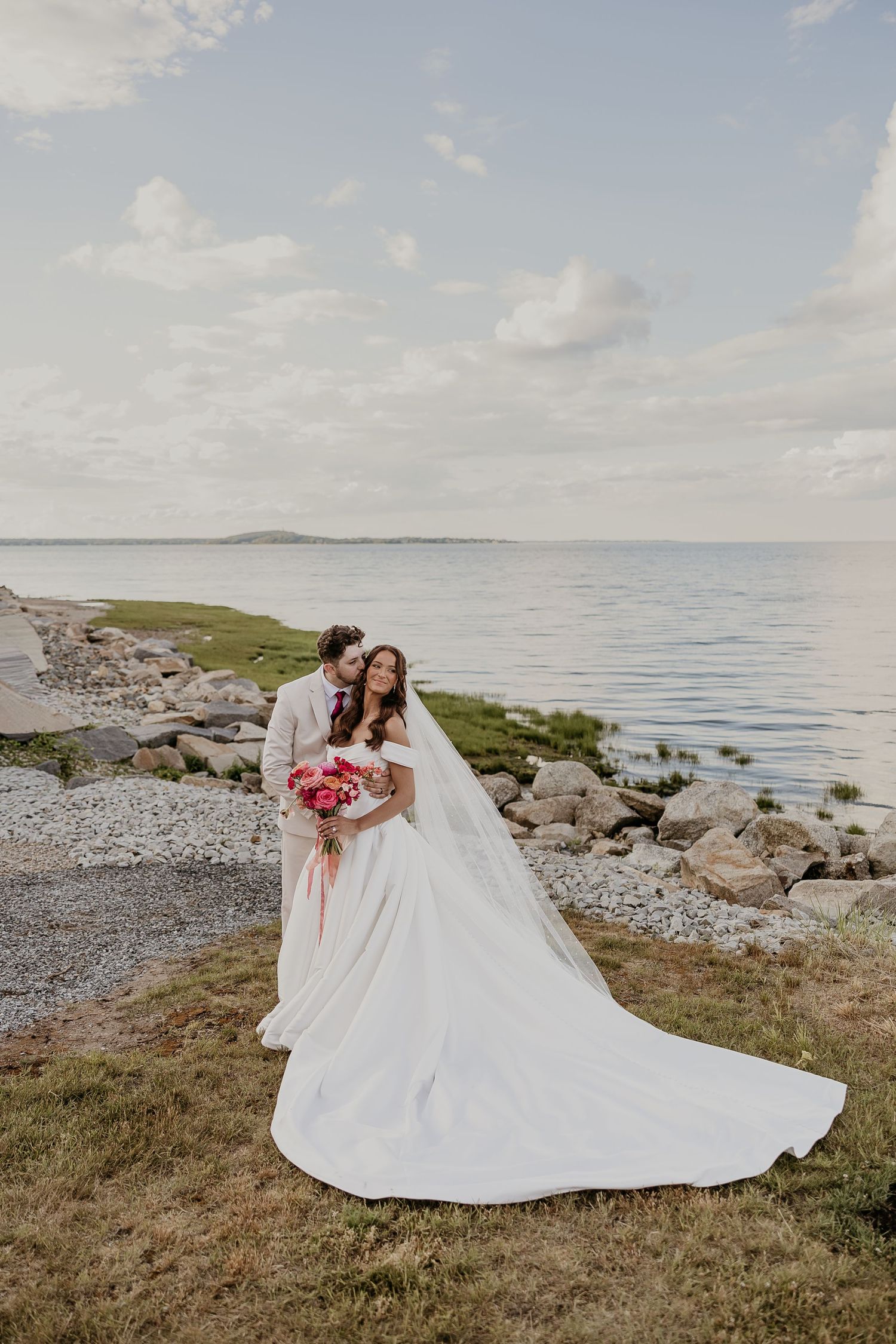 Bride and groom pose by rocky seaside, her white gown trailing on grass, holding pink bouquet under cloudy sky.