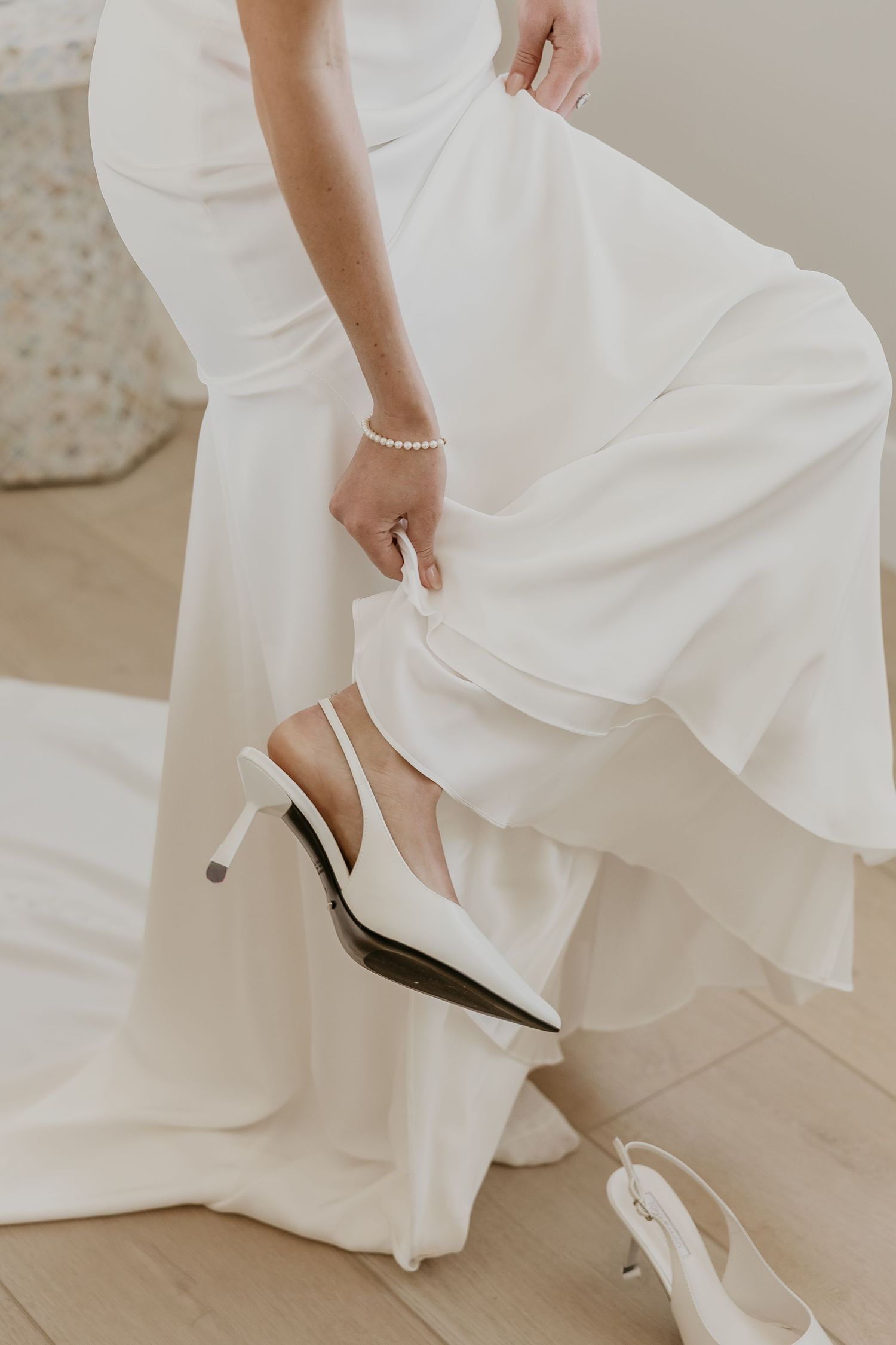 Bride in a white satin wedding gown putting on white slingback heels on a wooden floor.