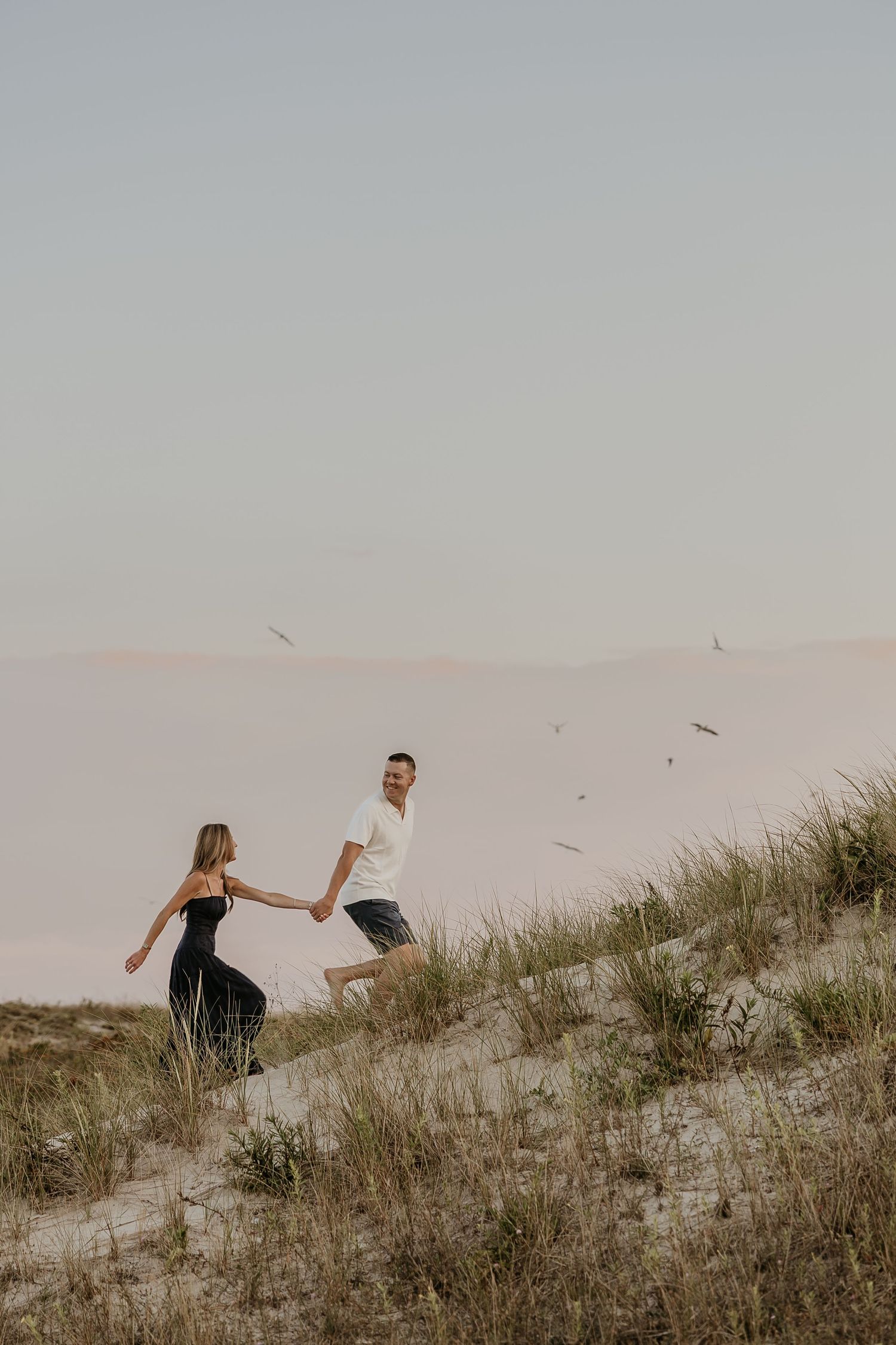 A couple holds hands while running up a sandy dune with beach grass, the woman in black dress, man in white shirt.