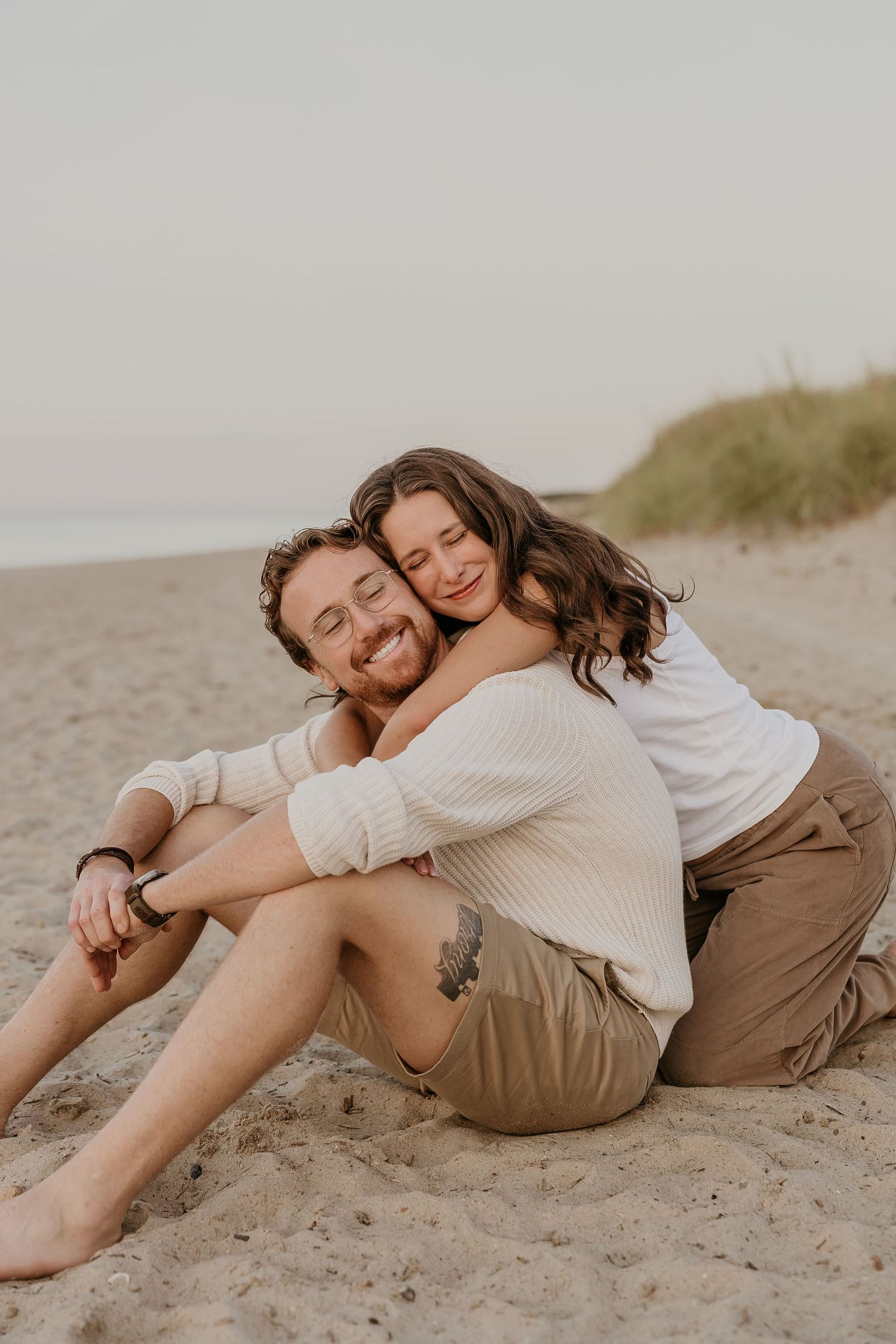 A happy couple embraces on a sandy beach, the woman hugging the man from behind as they smile joyfully.