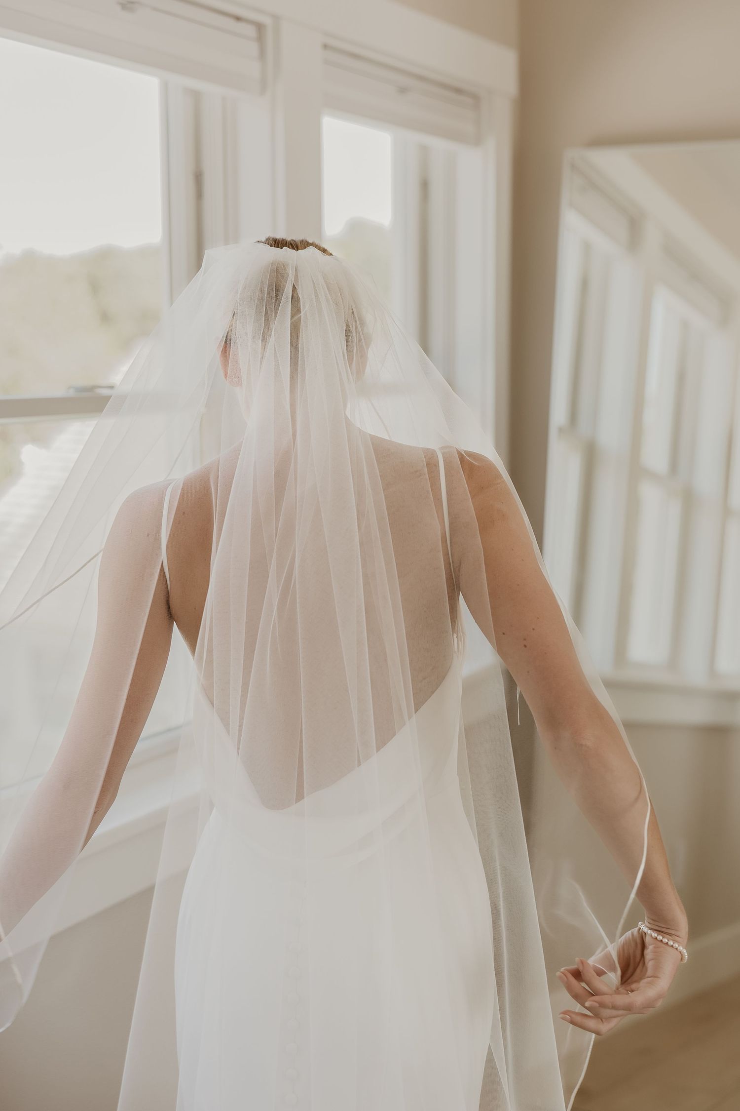 Bride in white gown and flowing veil seen from behind near bright windows.