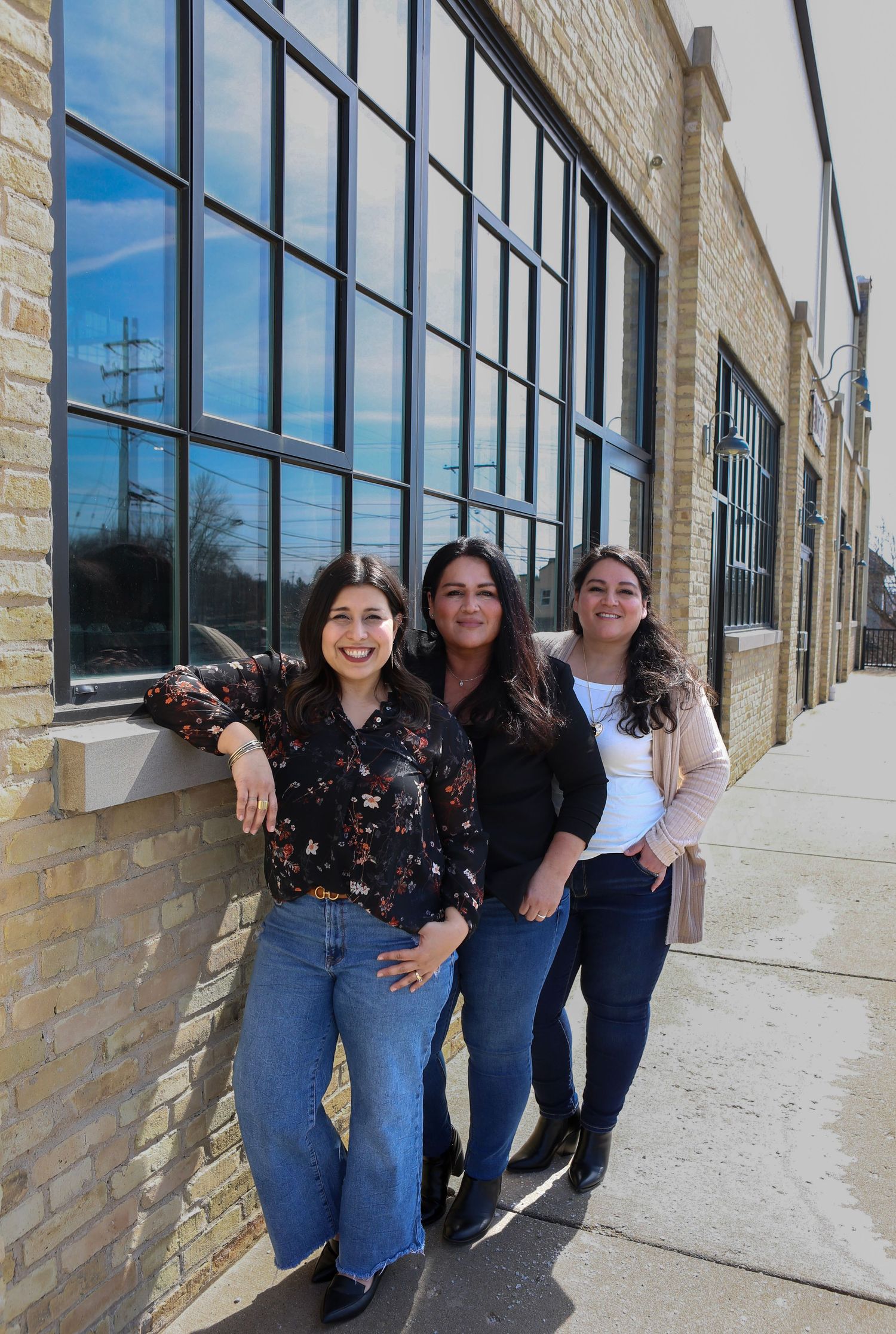 A group of people in casual clothing lean against a modern building's exterior wall with large windows.