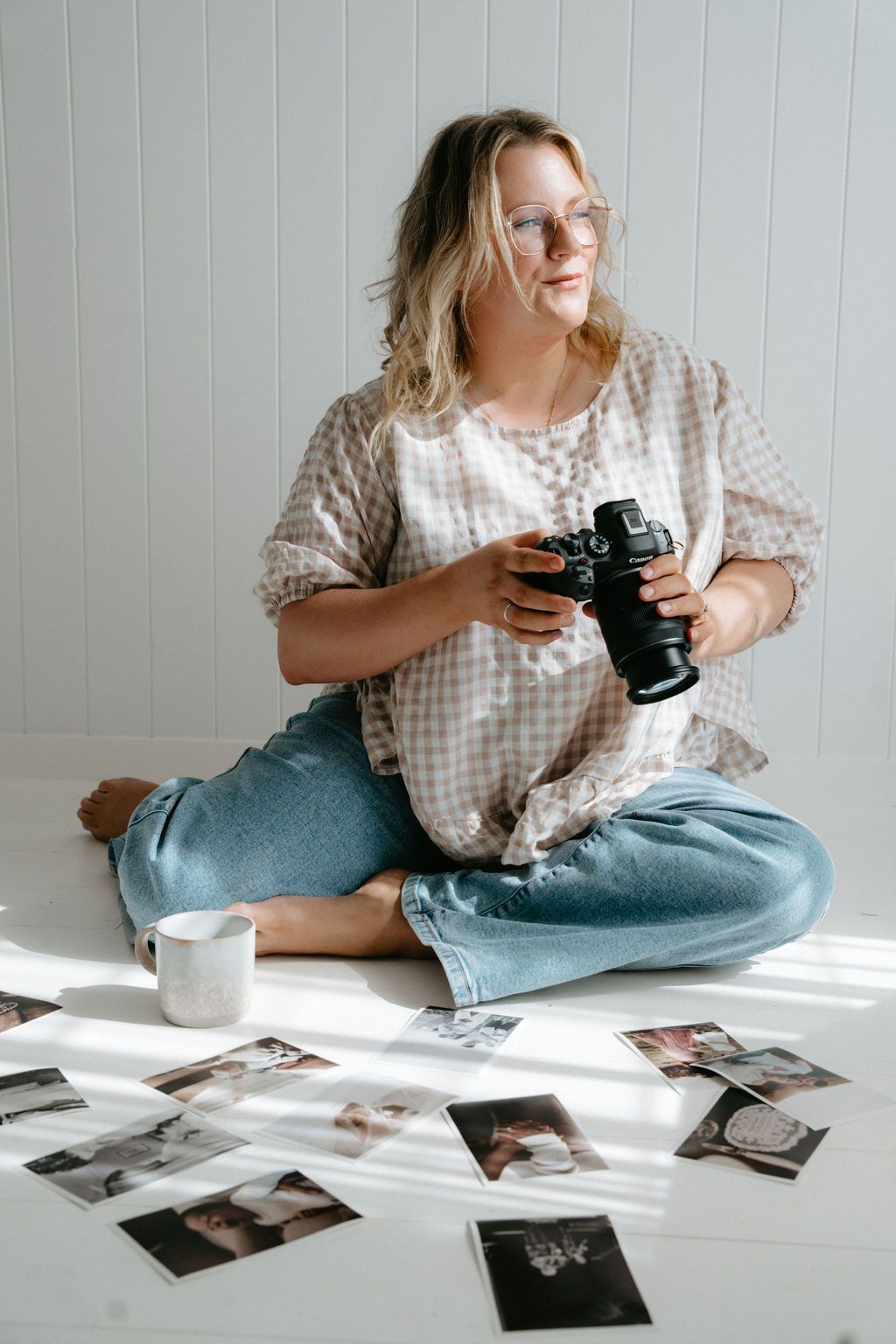 Photographer sitting cross-legged on white floor reviewing photos with camera in hand and prints spread around.
